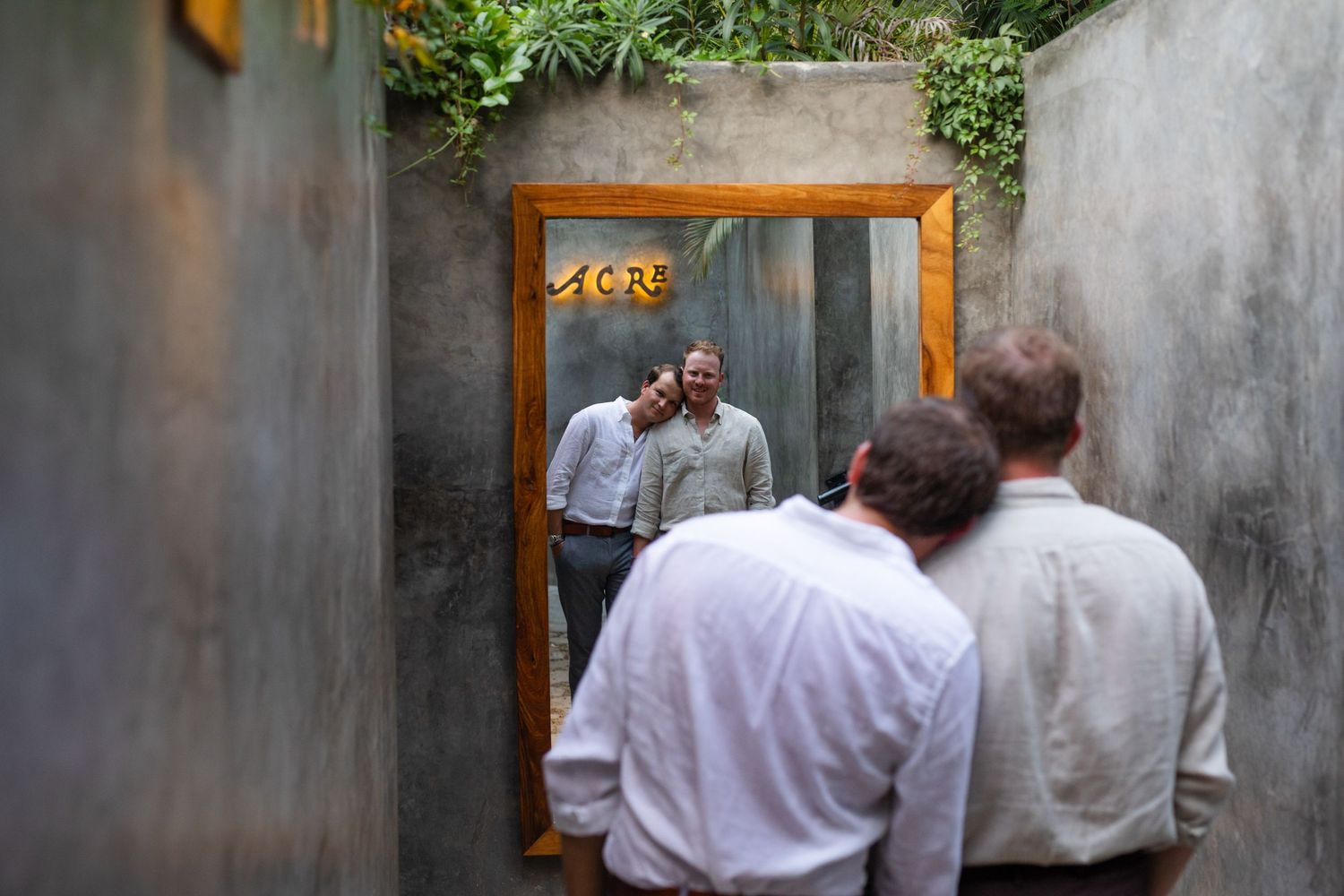 Two men in white shirts view their reflection in a wooden-framed mirror outdoors near a concrete wall at acre resort