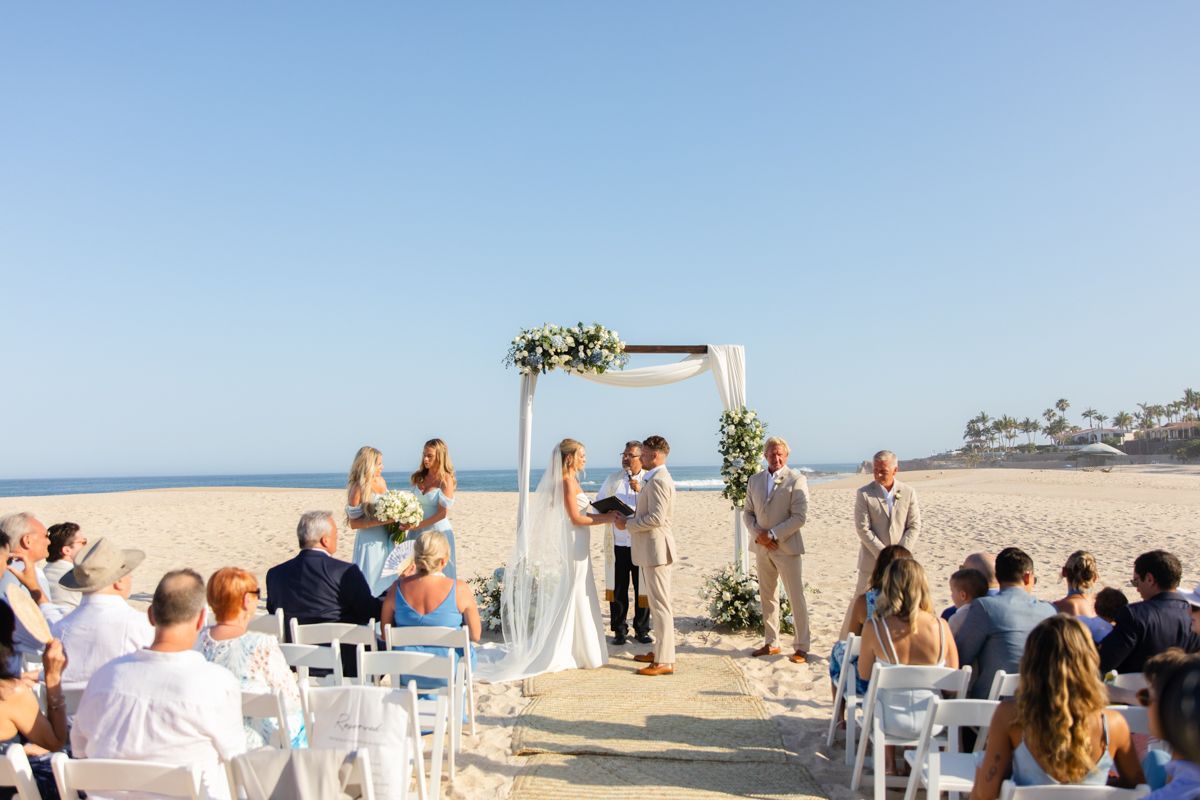 Beach wedding ceremony with couple at floral arch, guests seated on white chairs in sand.
