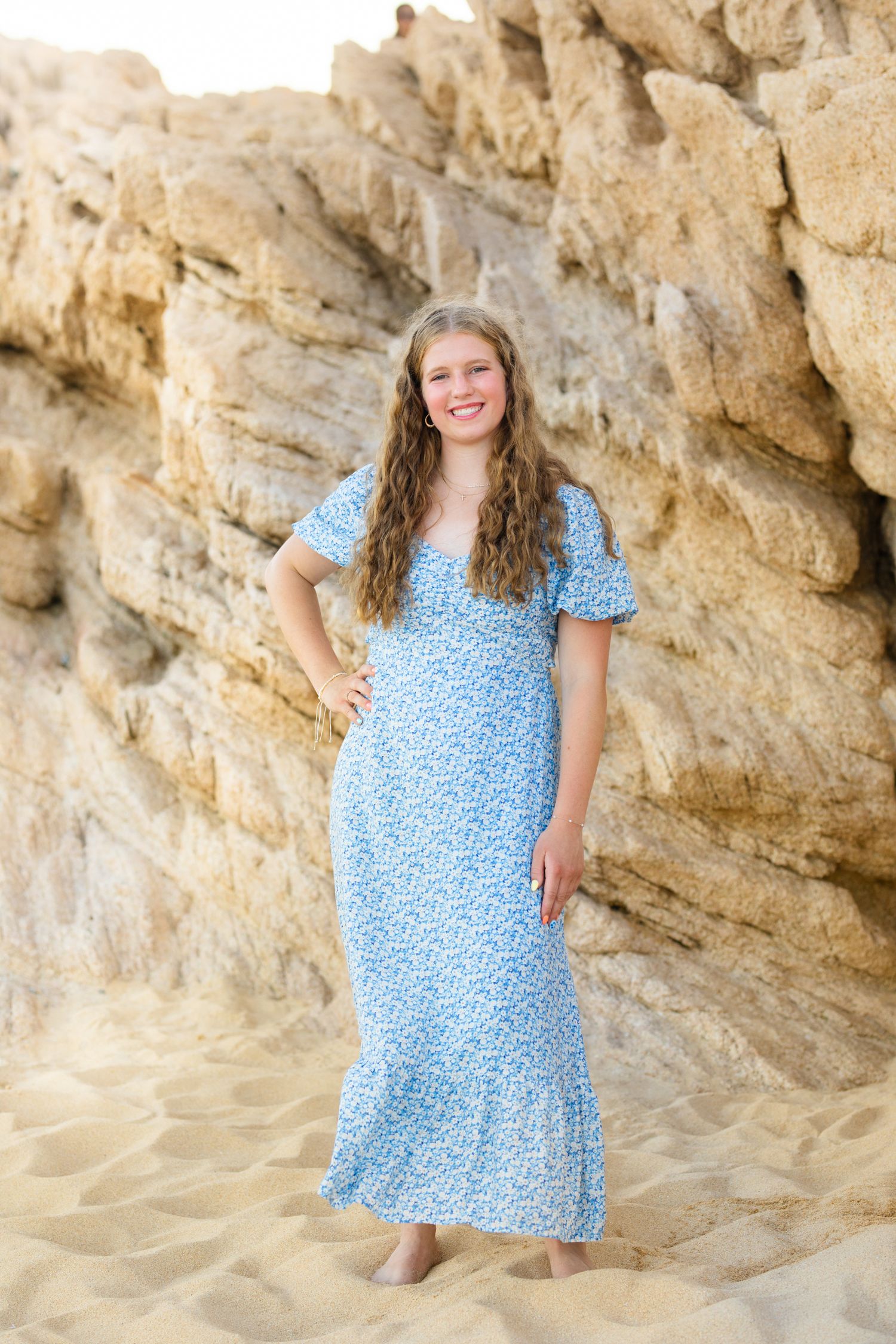 Curly-haired teen in blue floral maxi dress standing barefoot on sand near rocky cliff wall.
