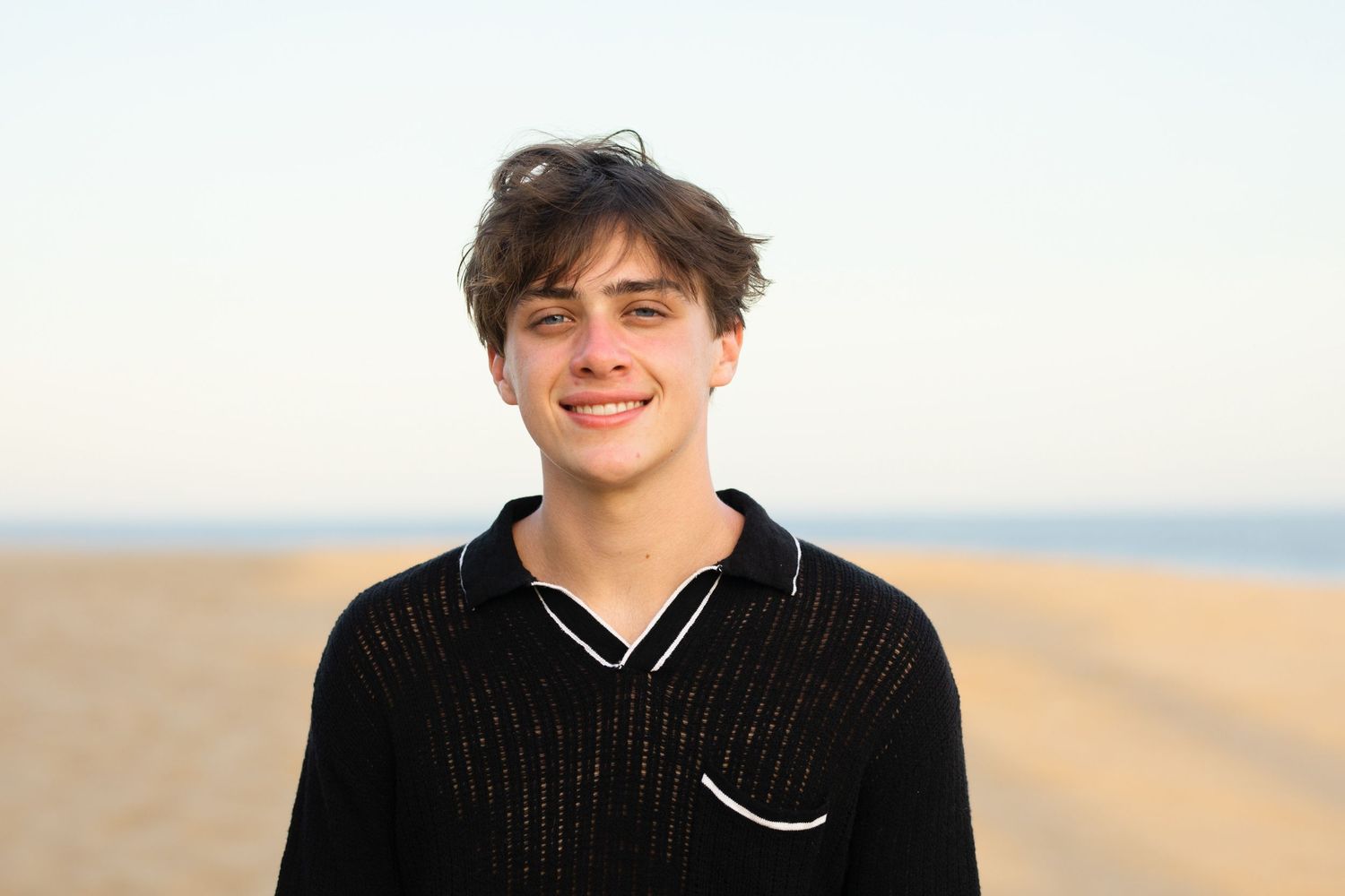 Smiling teenage boy in black polo shirt posing on sandy beach at golden hour.