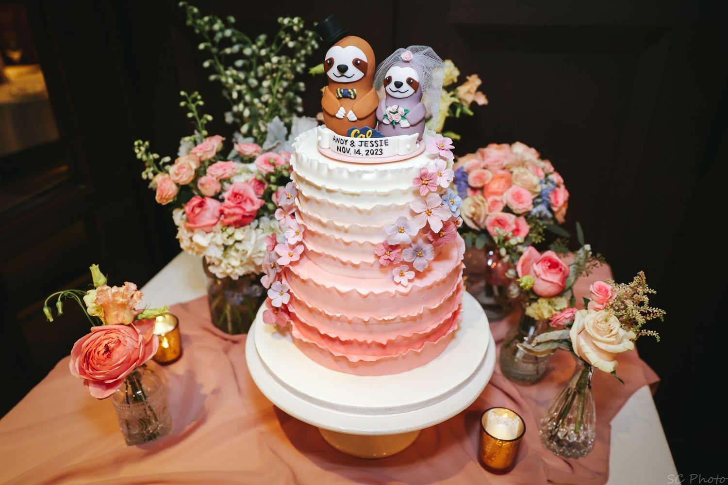 A pink wedding cake decorated with floral details and owl toppers on display with candles.
