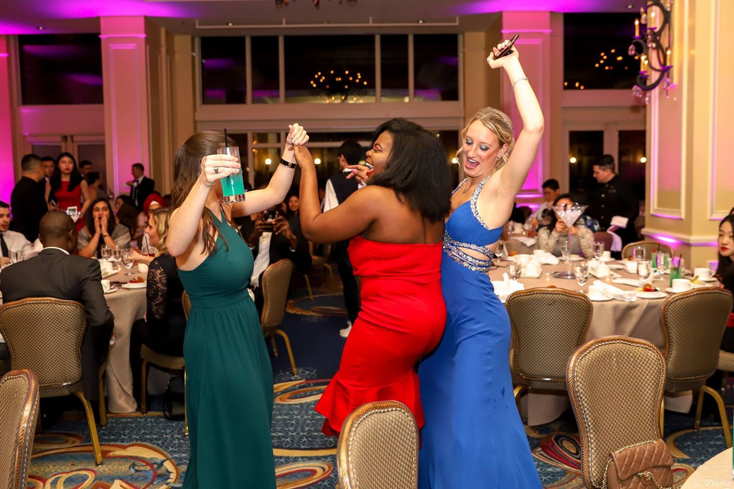 Three women in formal dresses dancing and celebrating at an elegant event.