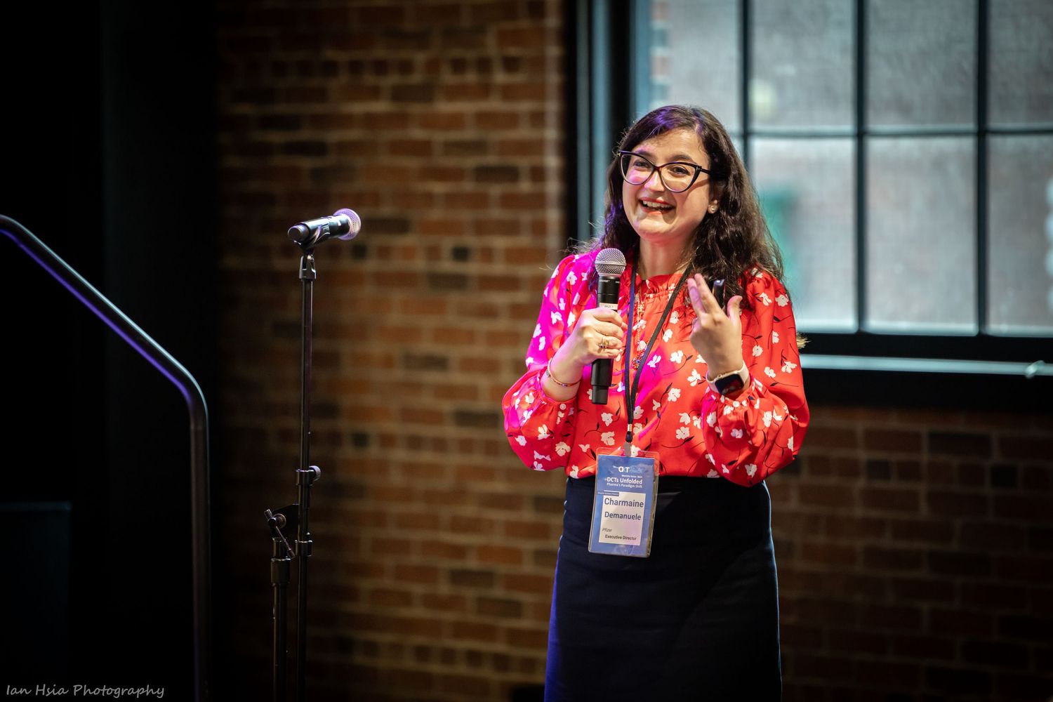 Speaker in bright pink polka dot top presenting at event with brick wall background.
