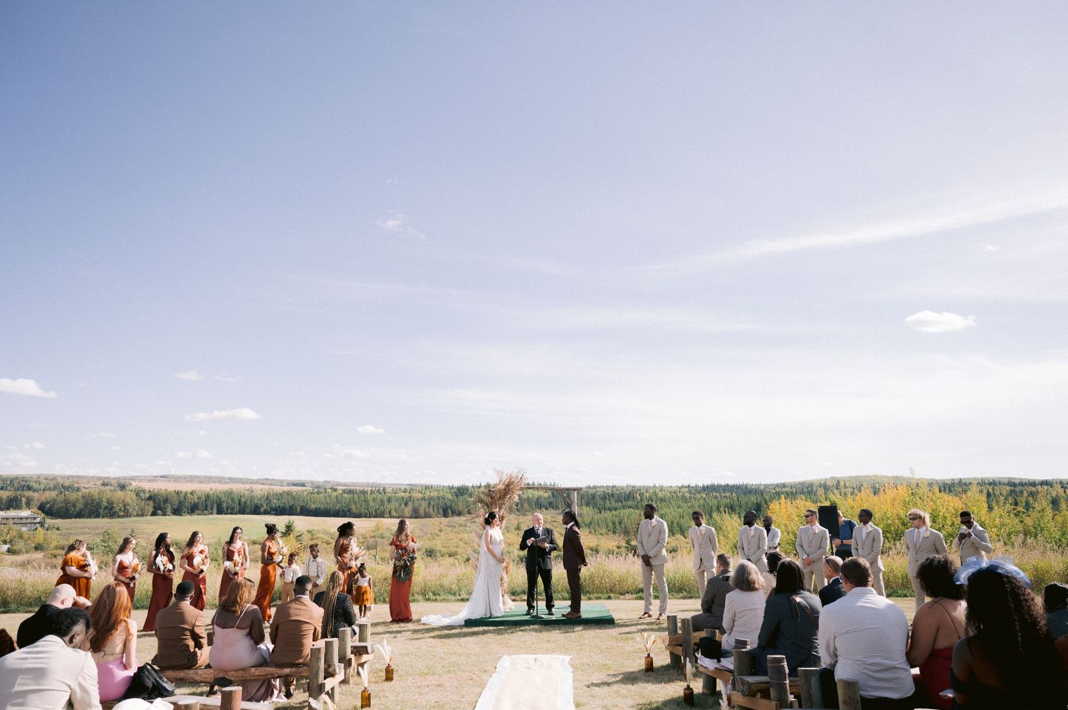 A wide panoramic view of an outdoor wedding ceremony with guests seated in an open field.