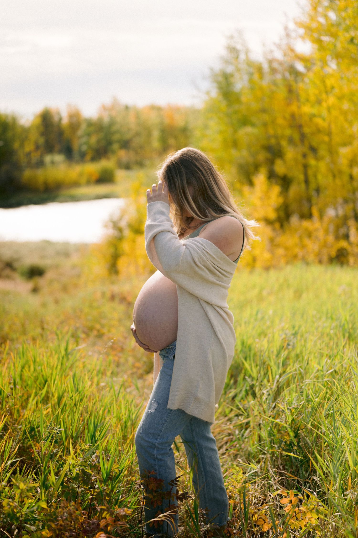 A pregnant woman stands in a meadow near a lake during autumn, wearing jeans and a cream sweater amidst golden foliage.