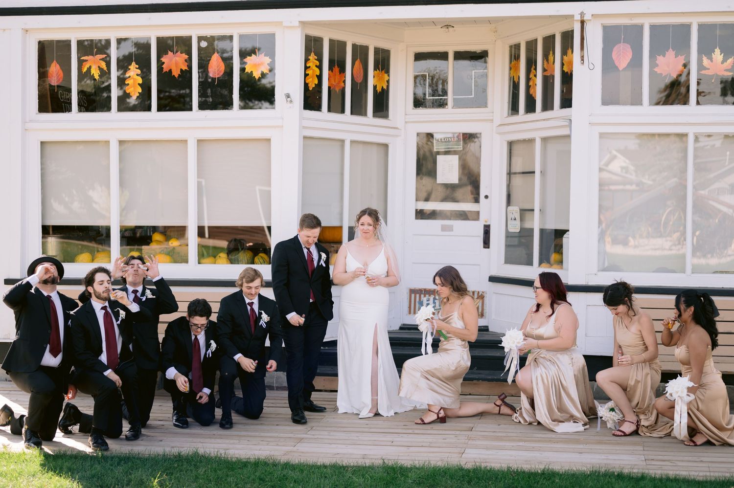 Wedding party members in black suits and champagne dresses sit together on a white porch during an outdoor celebration.