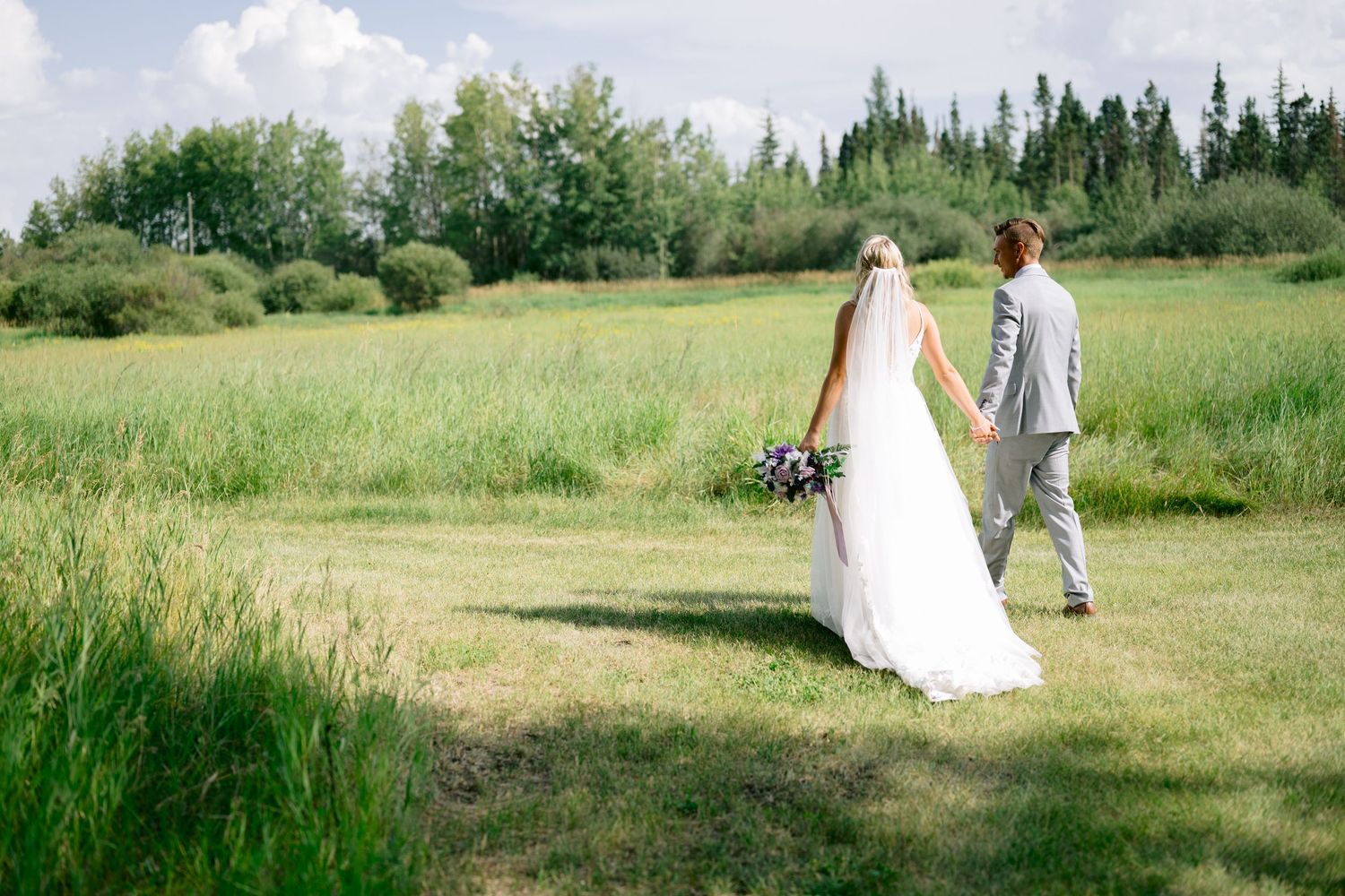 Bride and groom holding hands walking through a lush green field on their wedding day.