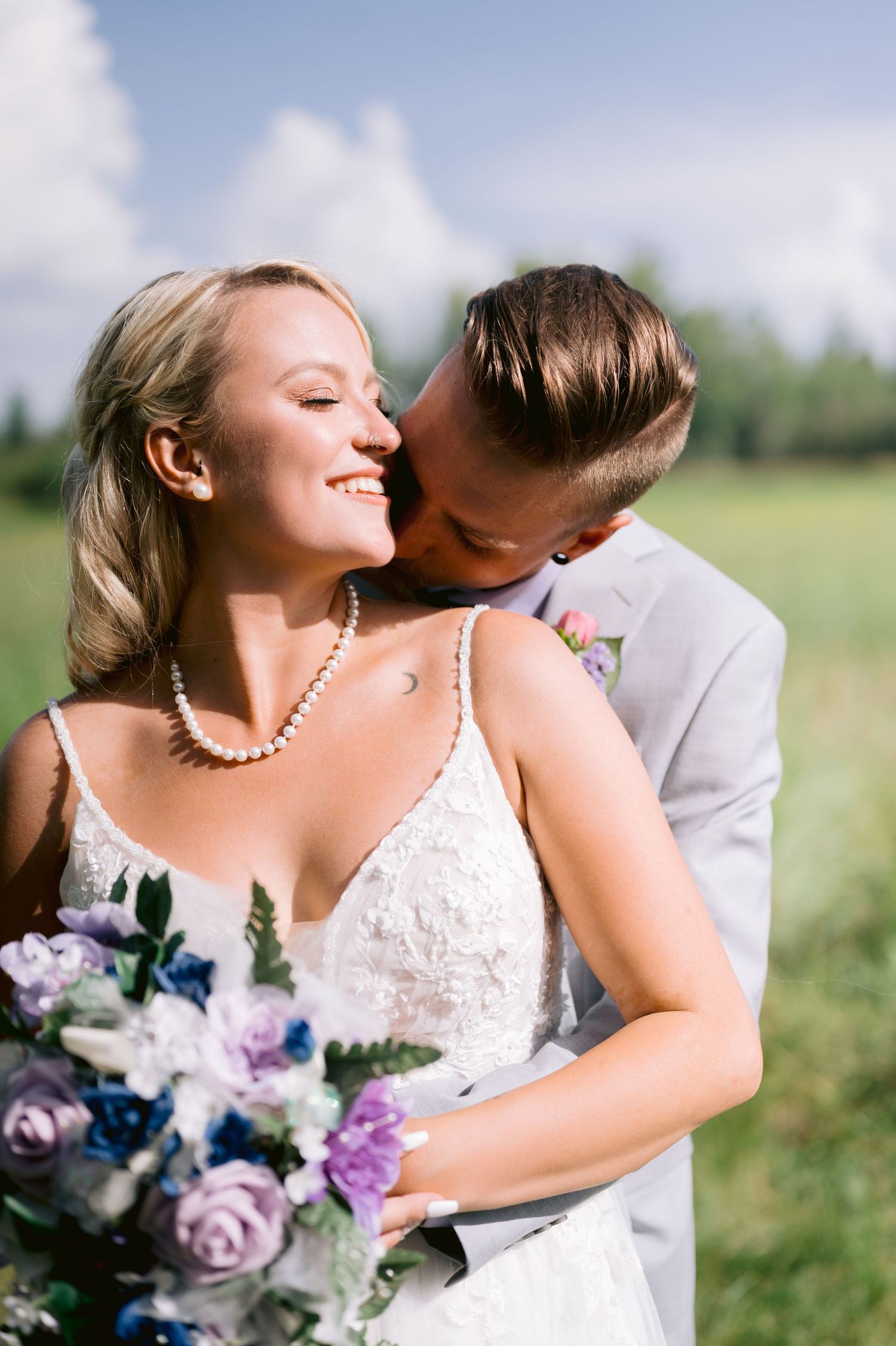 A romantic wedding couple shares intimate moments in a sunlit field with purple and blue floral bouquet details.