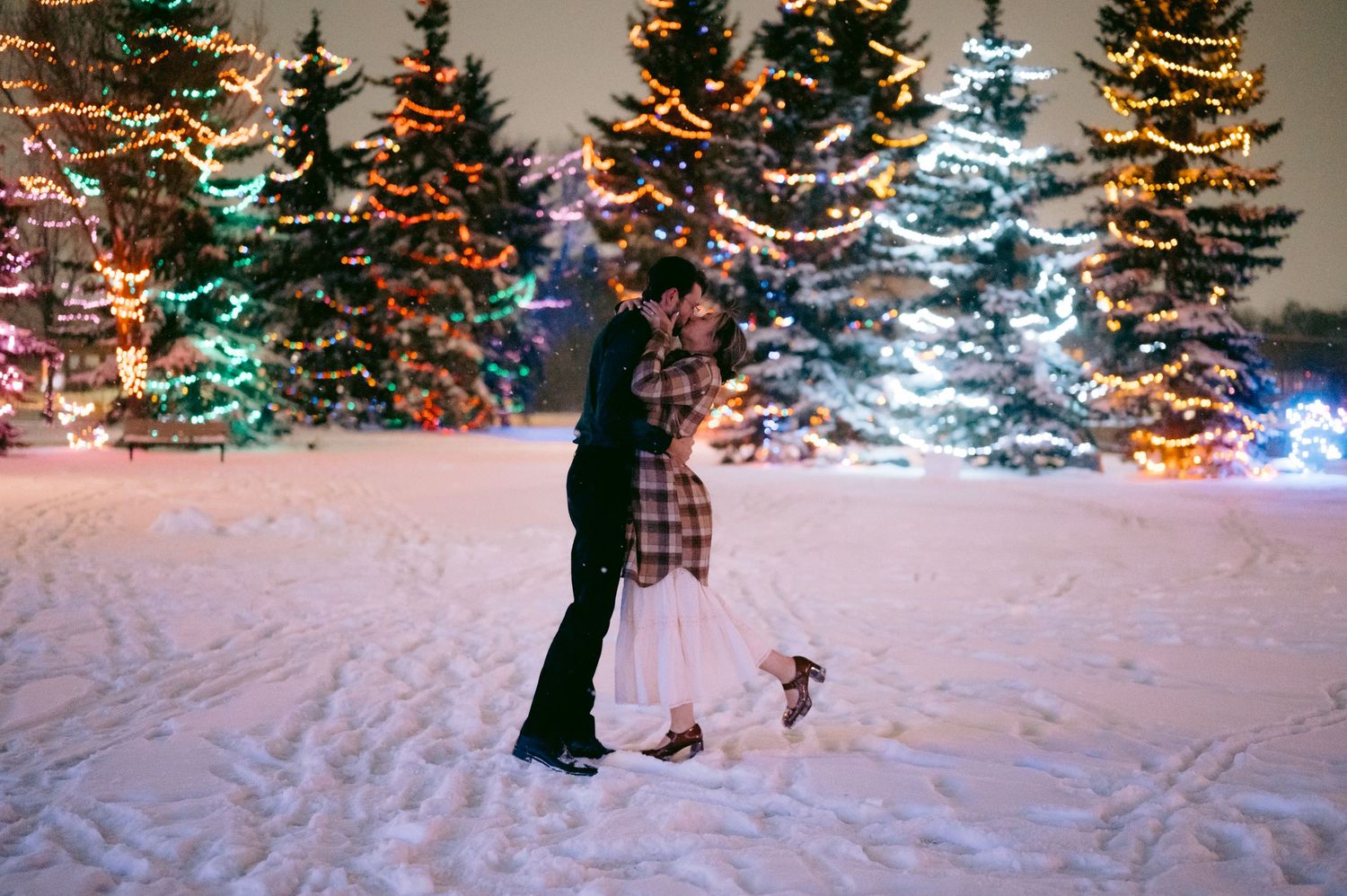 People walk through a snowy winter scene with illuminated Christmas trees glowing in the background.