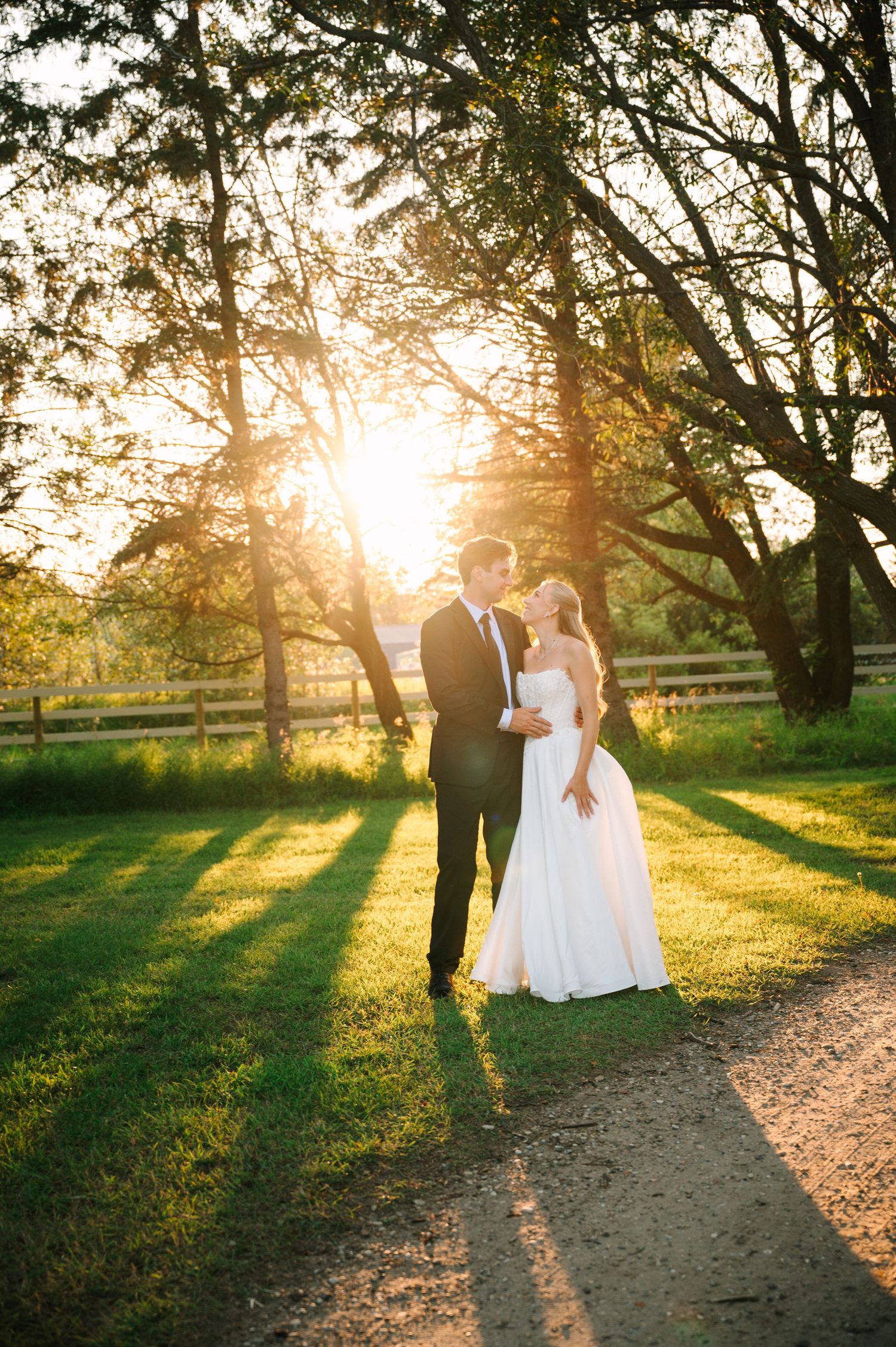 Bride and groom share a romantic moment at golden sunset, standing on a path surrounded by lush green trees.