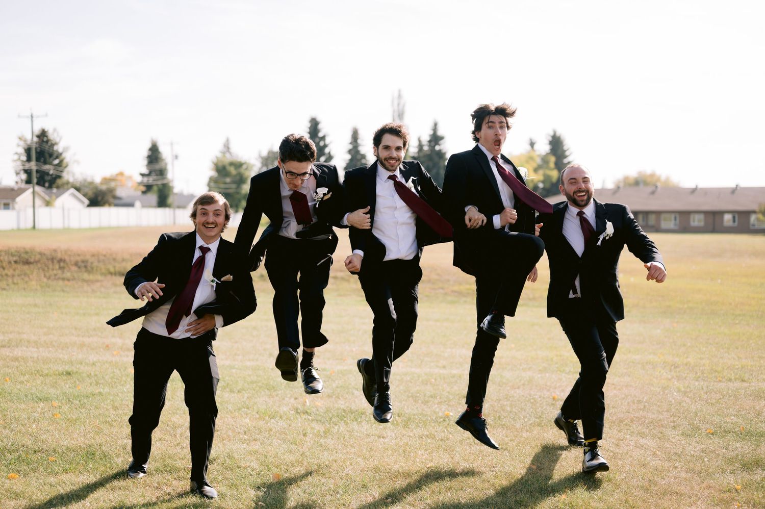 Groomsmen in black suits jump playfully on a grassy field during a wedding photo session.