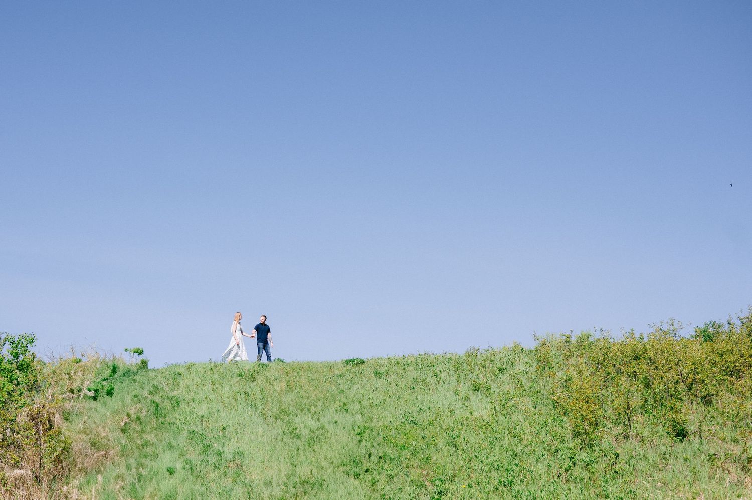 Two small figures silhouetted on a grassy hilltop against a bright blue sky on a sunny day.