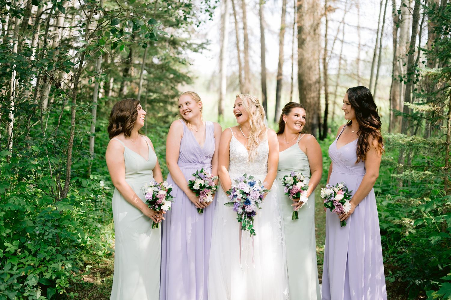 Bridesmaids in lavender dresses stand together holding bouquets in a sunlit forest setting.