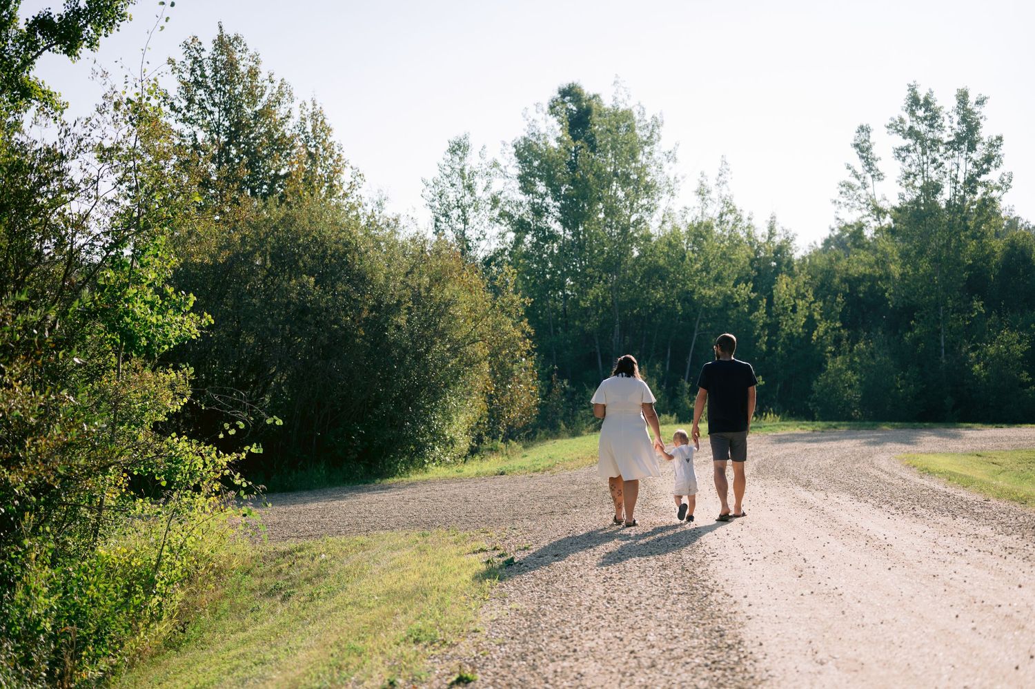 A family walks hand-in-hand down a sunny gravel path surrounded by lush green trees.
