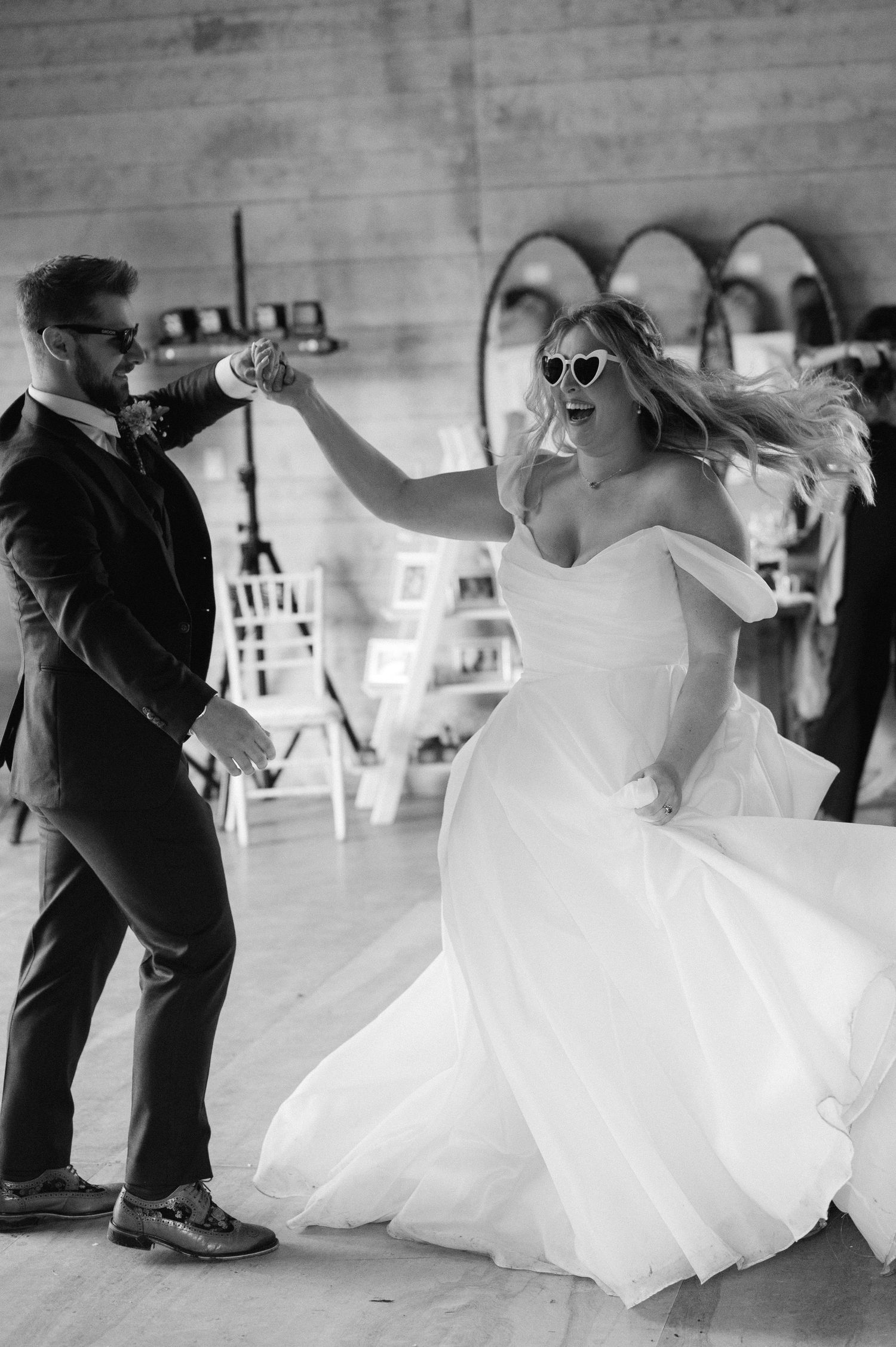 A couple dances joyfully at their wedding reception, with the bride in a flowing white dress and sunglasses.