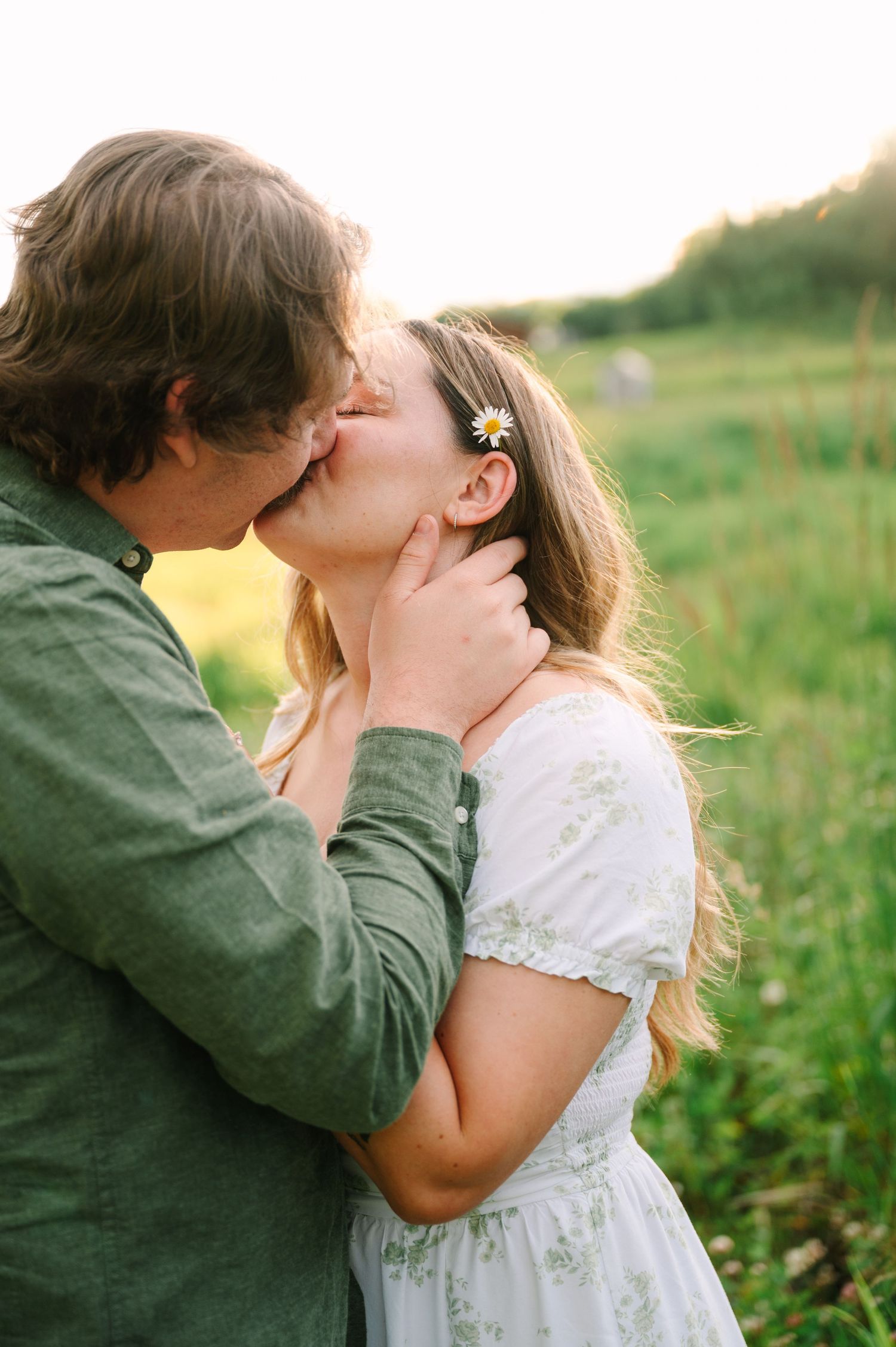 A couple shares a romantic kiss in a green field, the woman wearing a white floral dress with a daisy in her hair.