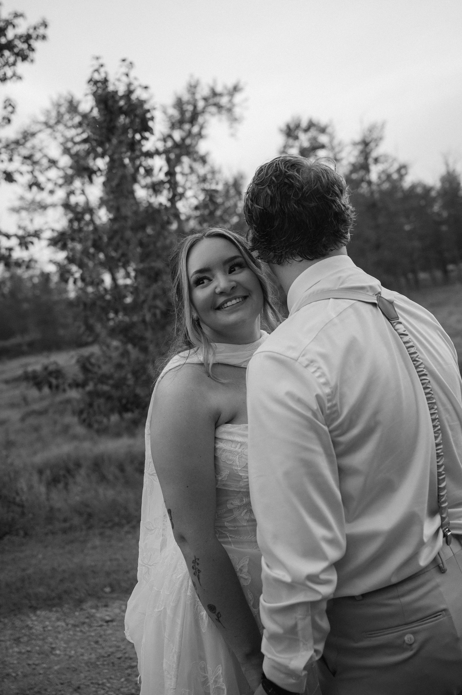 Black and white photo of a smiling bride in a white dress holding hands with a groom in suspenders outdoors.