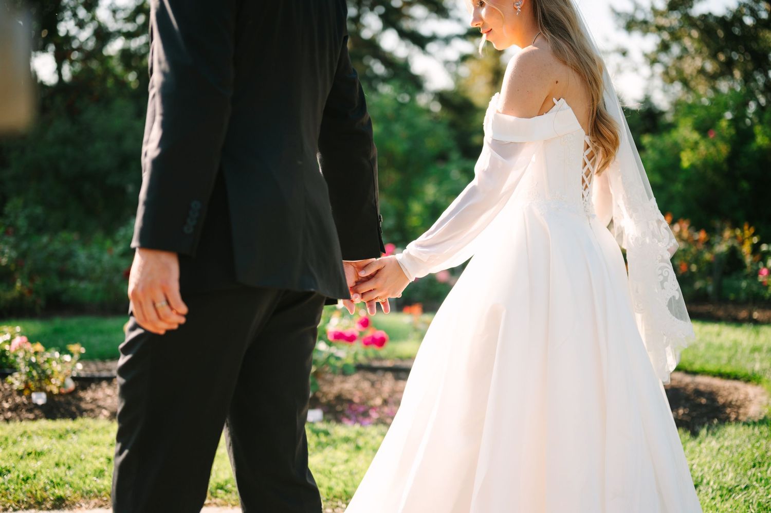 Bride in white off-shoulder gown holding hands with groom in black suit outdoors in garden setting.