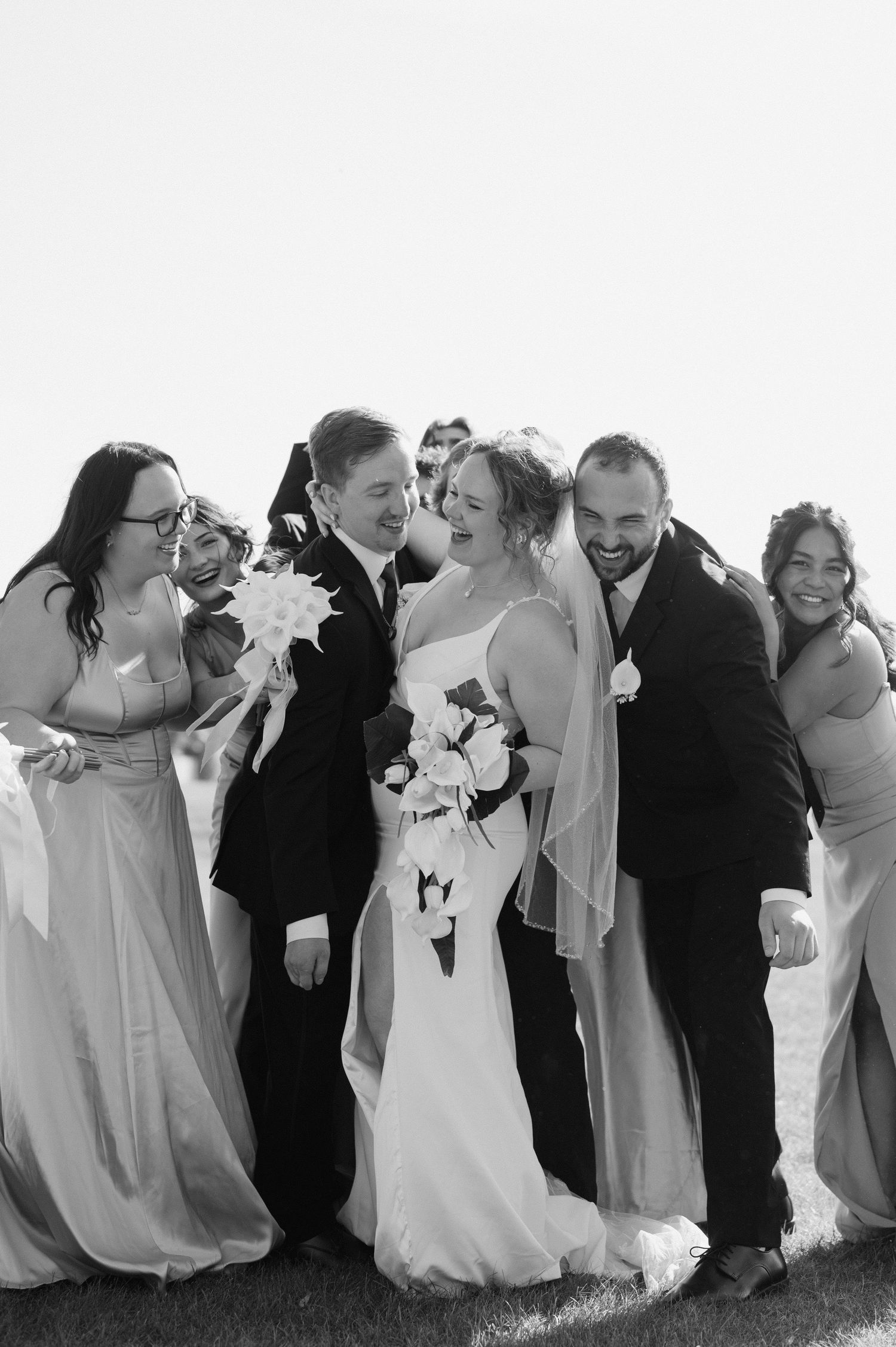 A joyful wedding party in black and white poses together outdoors, with bridesmaids in light dresses and groomsmen in dark suits.