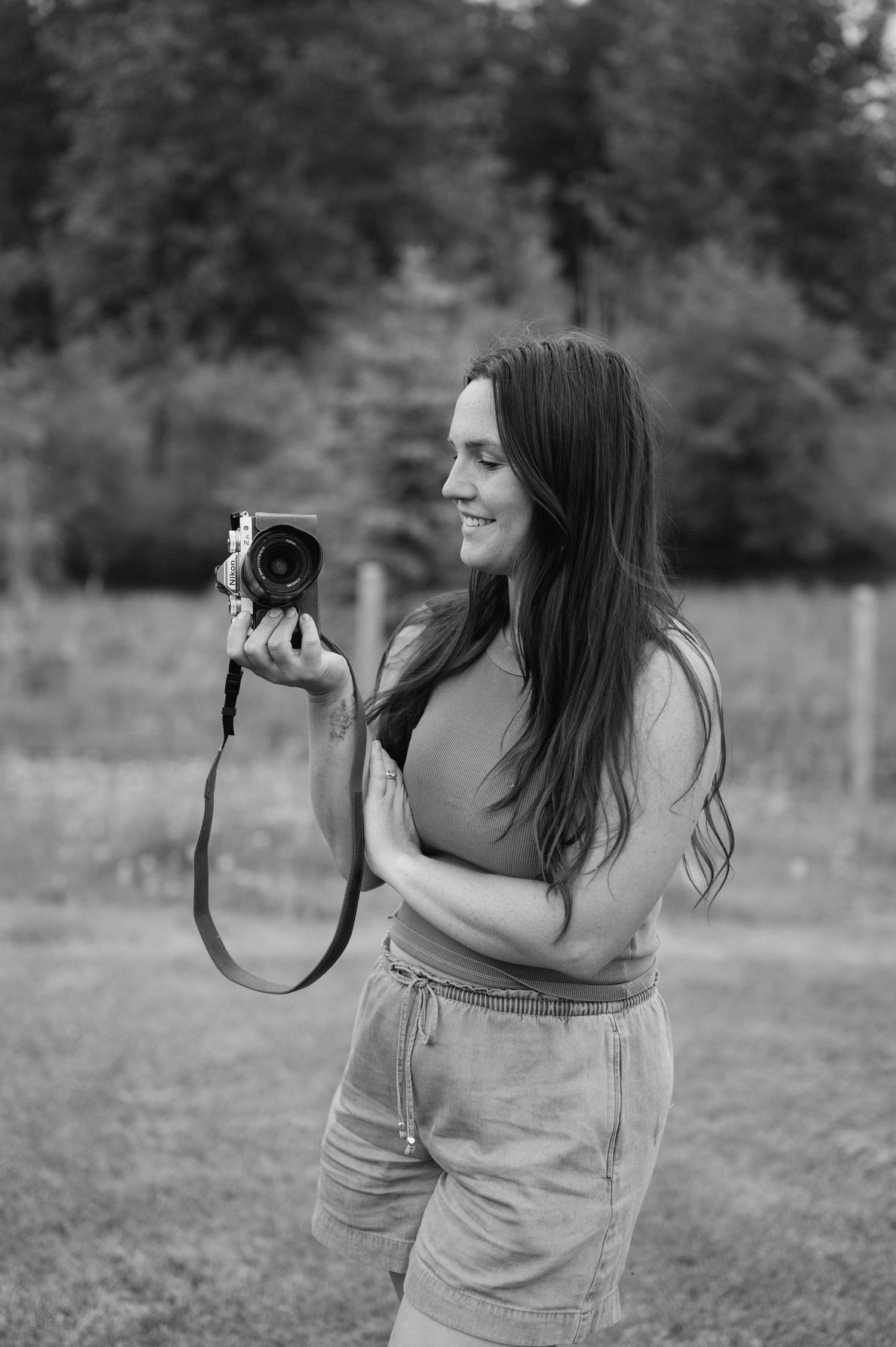 Black and white photo of a young woman with long hair holding a camera outdoors in a grassy field.