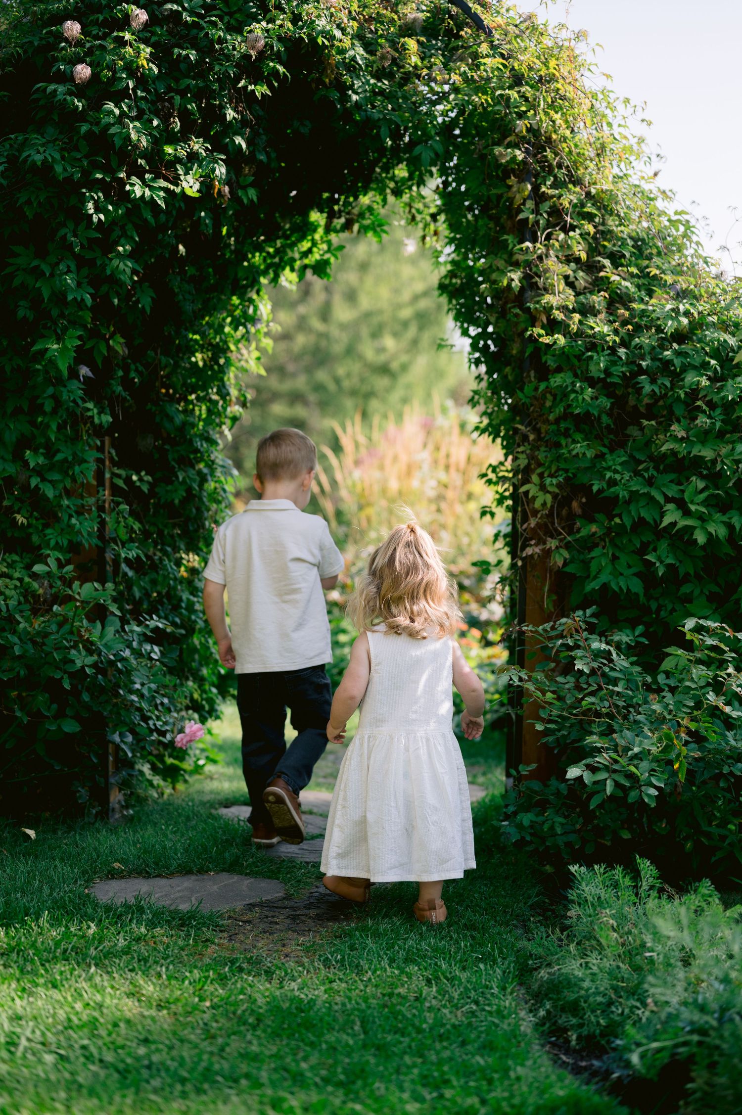 Two young children walk hand-in-hand through a lush green garden archway on a sunny day.
