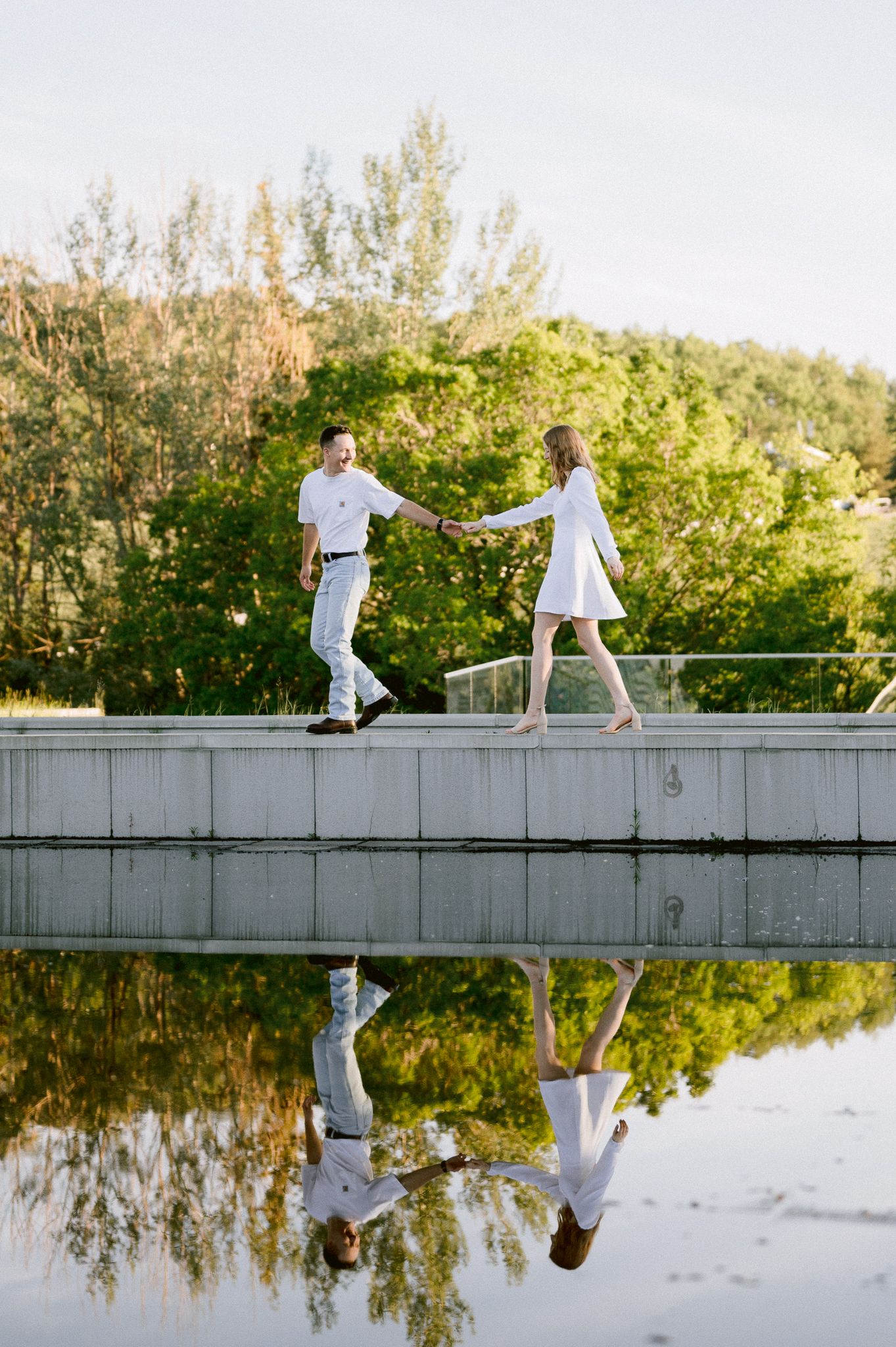 Two people in white outfits walking on a dock over still water creating mirror reflections at sunset.