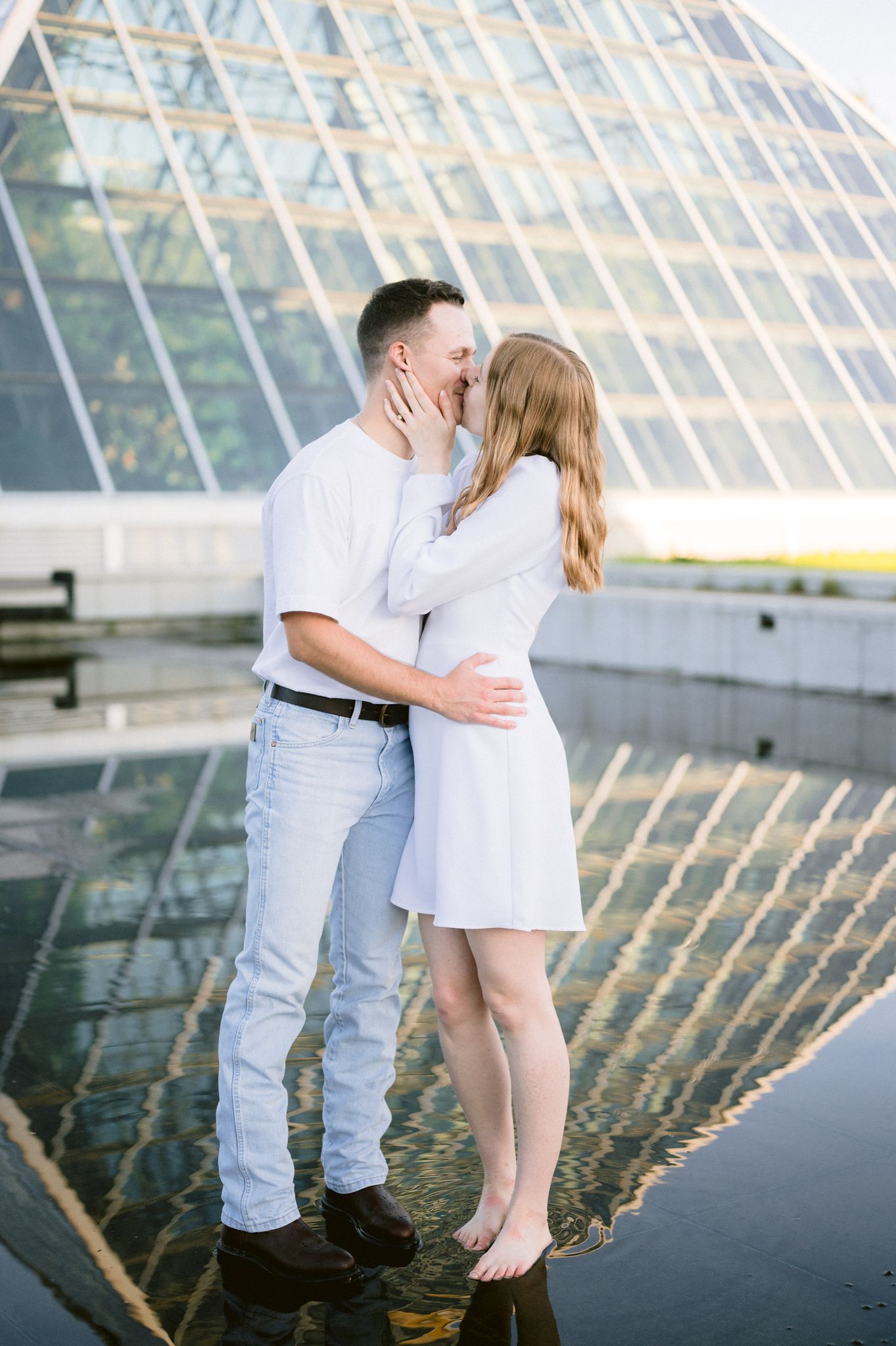 A couple in white and light colored clothing embraces near a modern glass building with reflective surfaces.