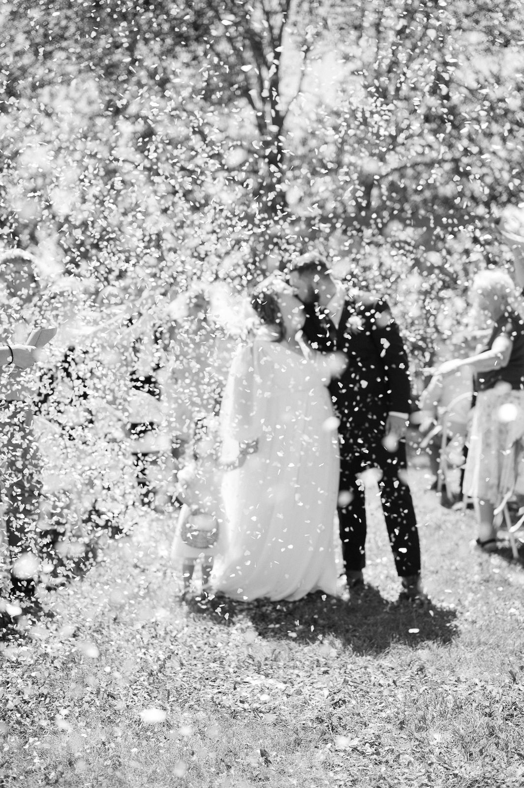 Black and white photo of a bride and groom kissing amid a shower of confetti at their outdoor wedding ceremony.