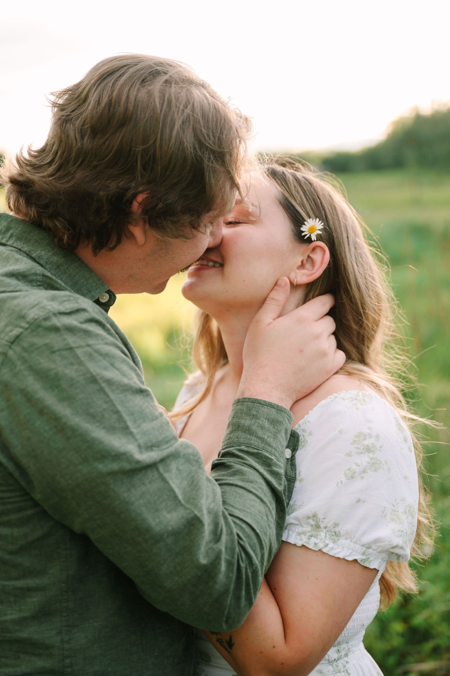 A couple sharing a tender kiss outdoors, the woman wearing a white floral dress with a daisy in her hair.