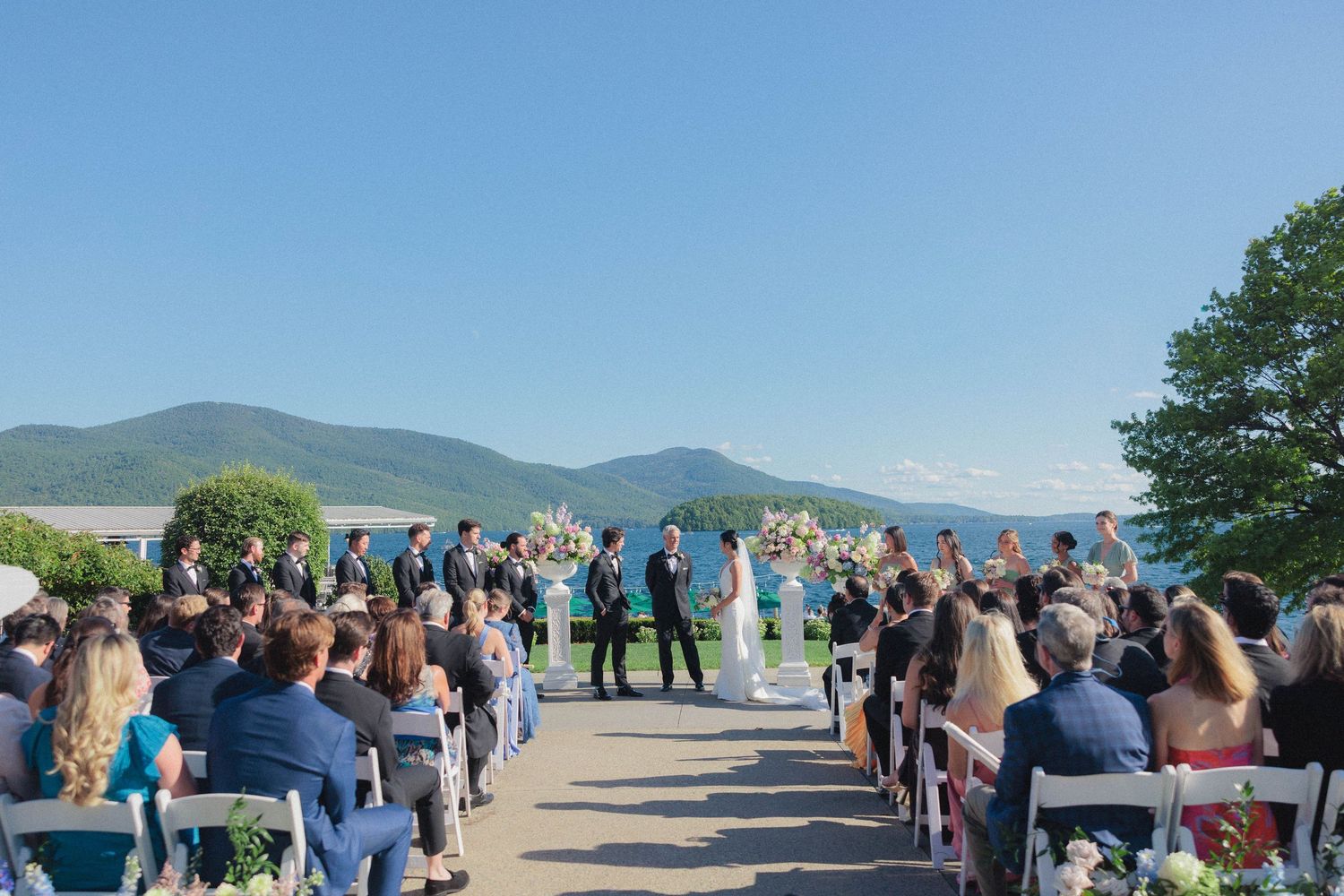 Outdoor wedding ceremony with mountain and lake backdrop under clear blue sky with seated guests.