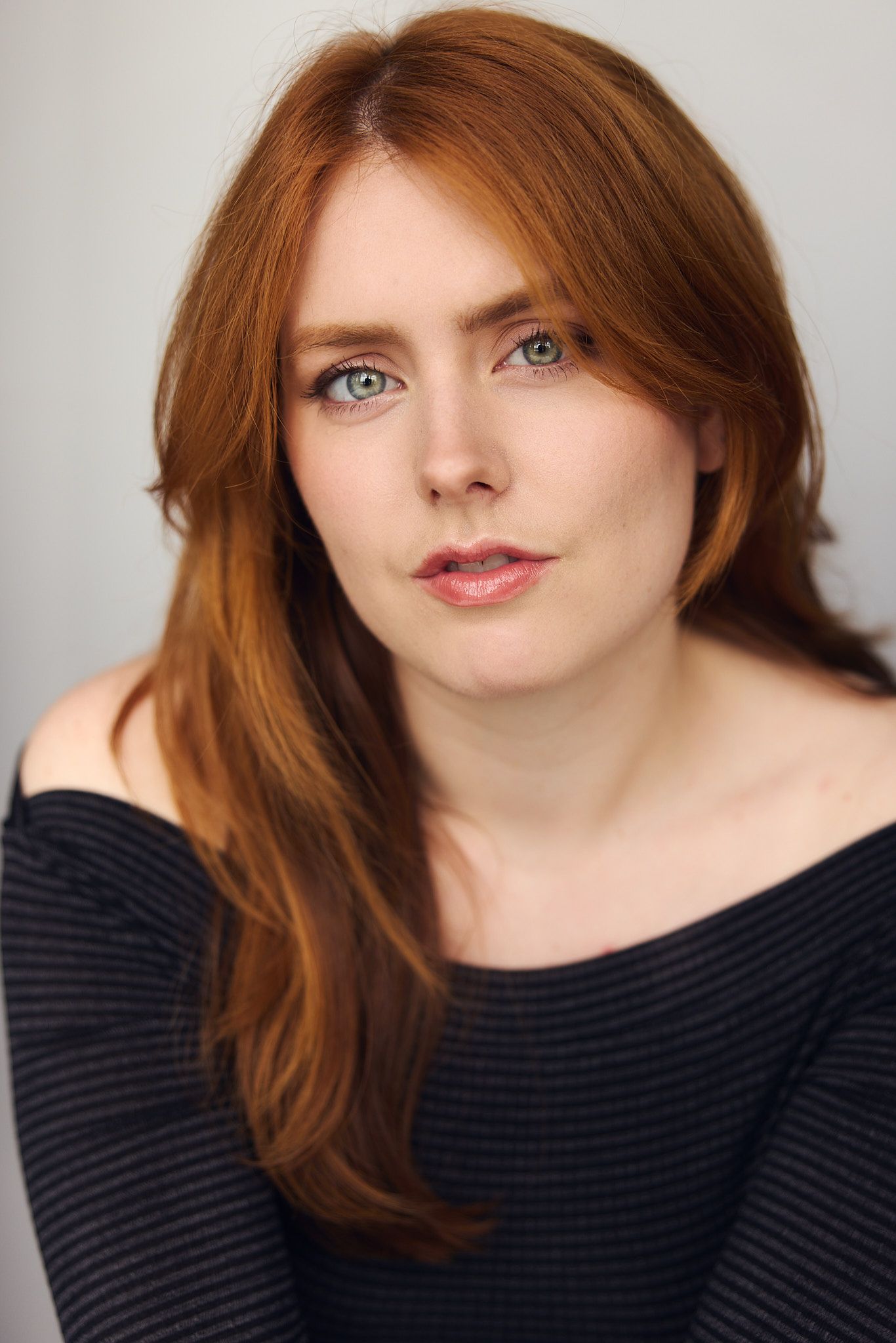 Young woman with long red hair and green eyes wearing an off-shoulder dark top in a professional headshot.