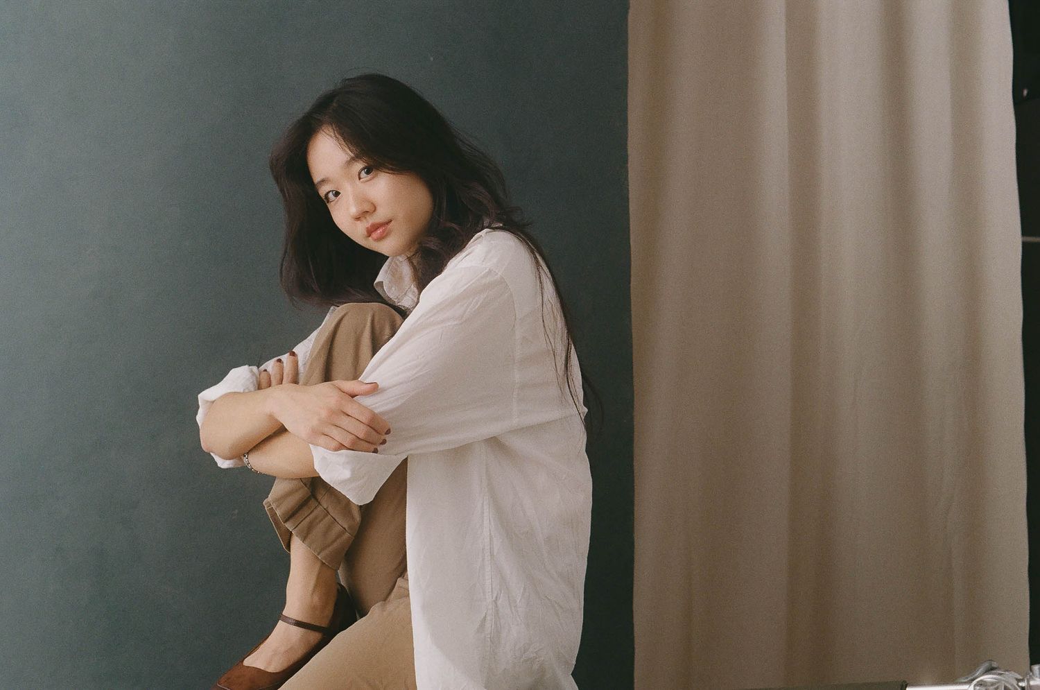 A person in a white sweater sits against a dark wall near curtains in a contemplative pose.