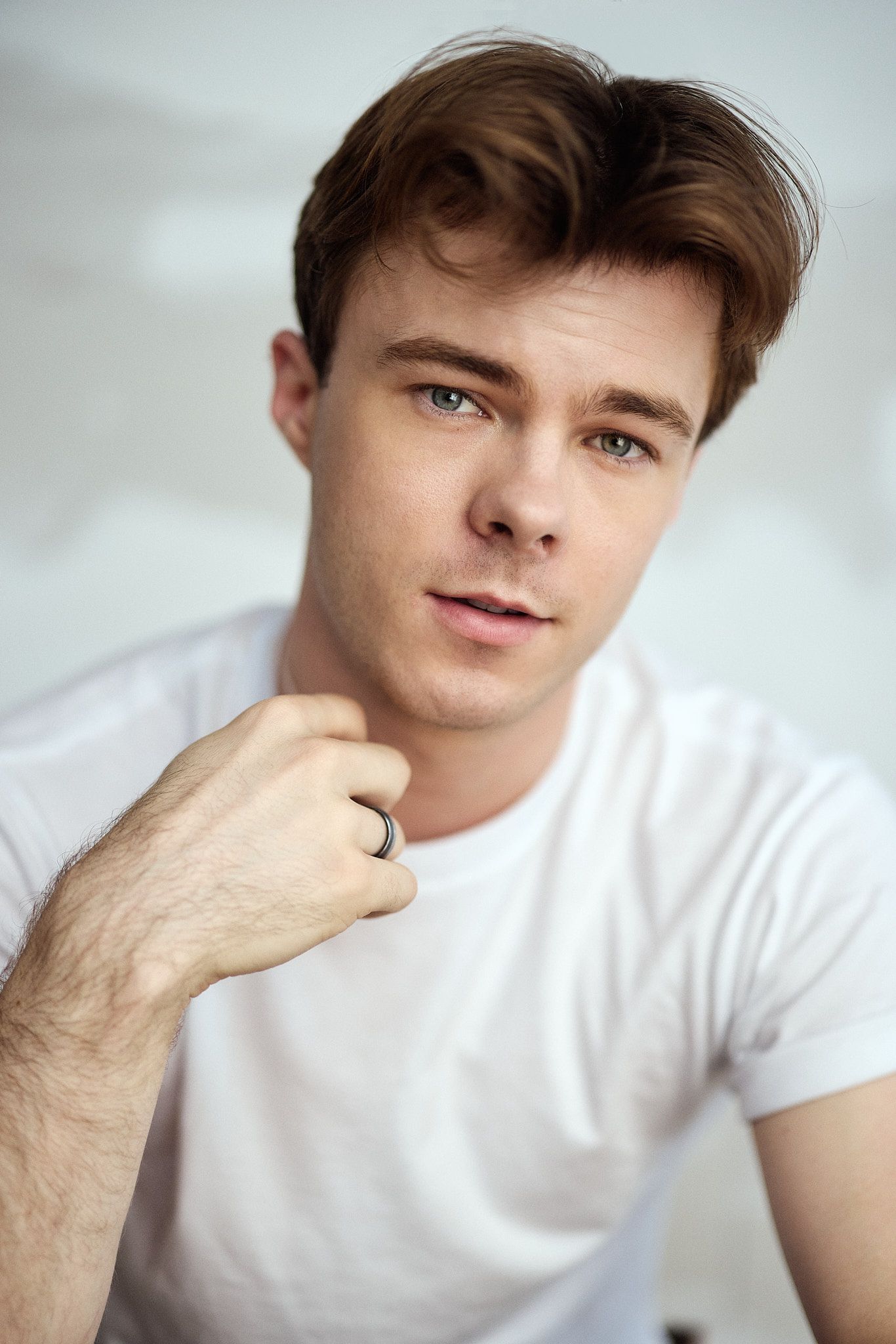 Young man with brown hair and blue eyes wearing a white t-shirt and silver ring, posing against a light background.