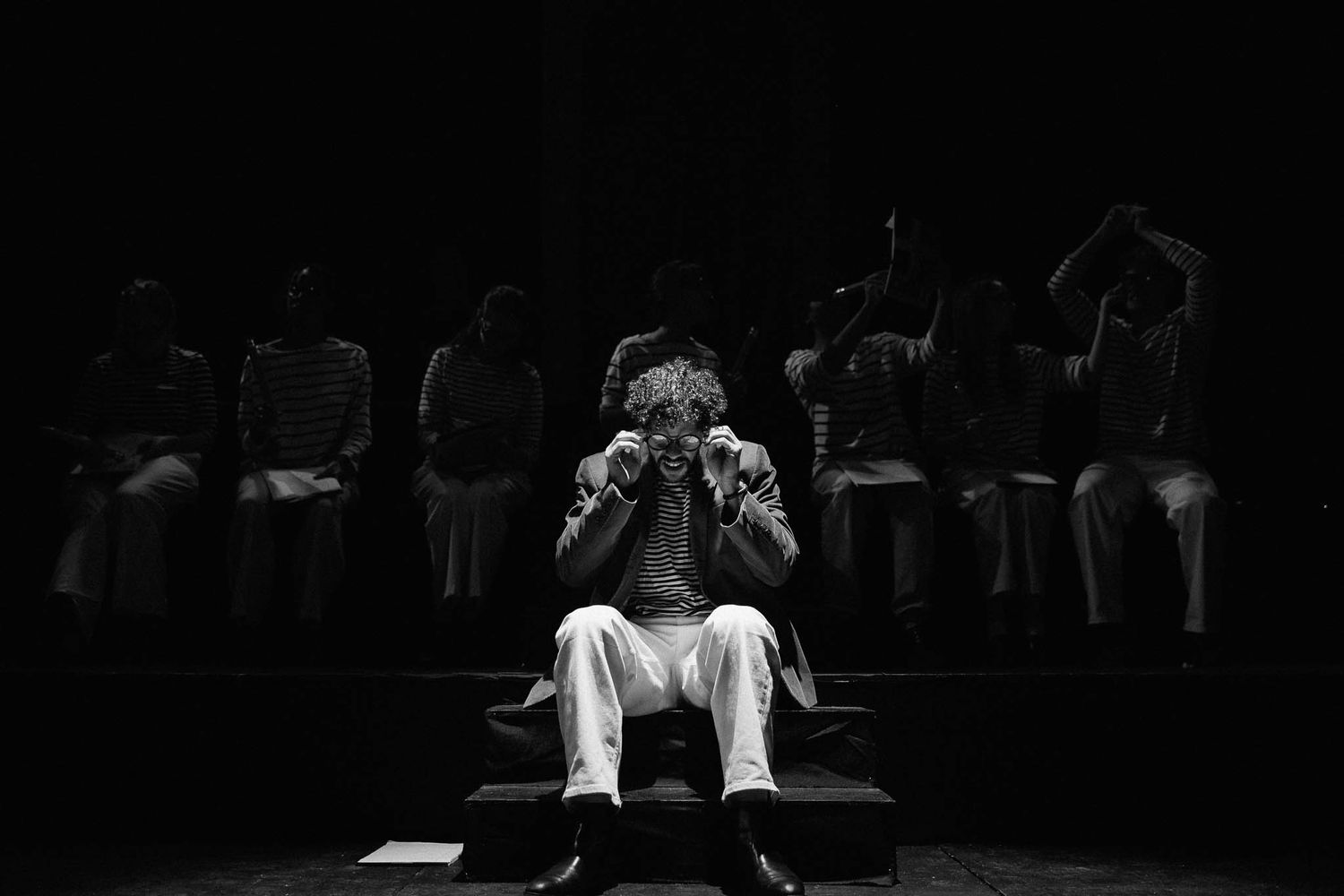 A dramatic black and white performance photo showing silhouetted figures on a dimly lit stage.