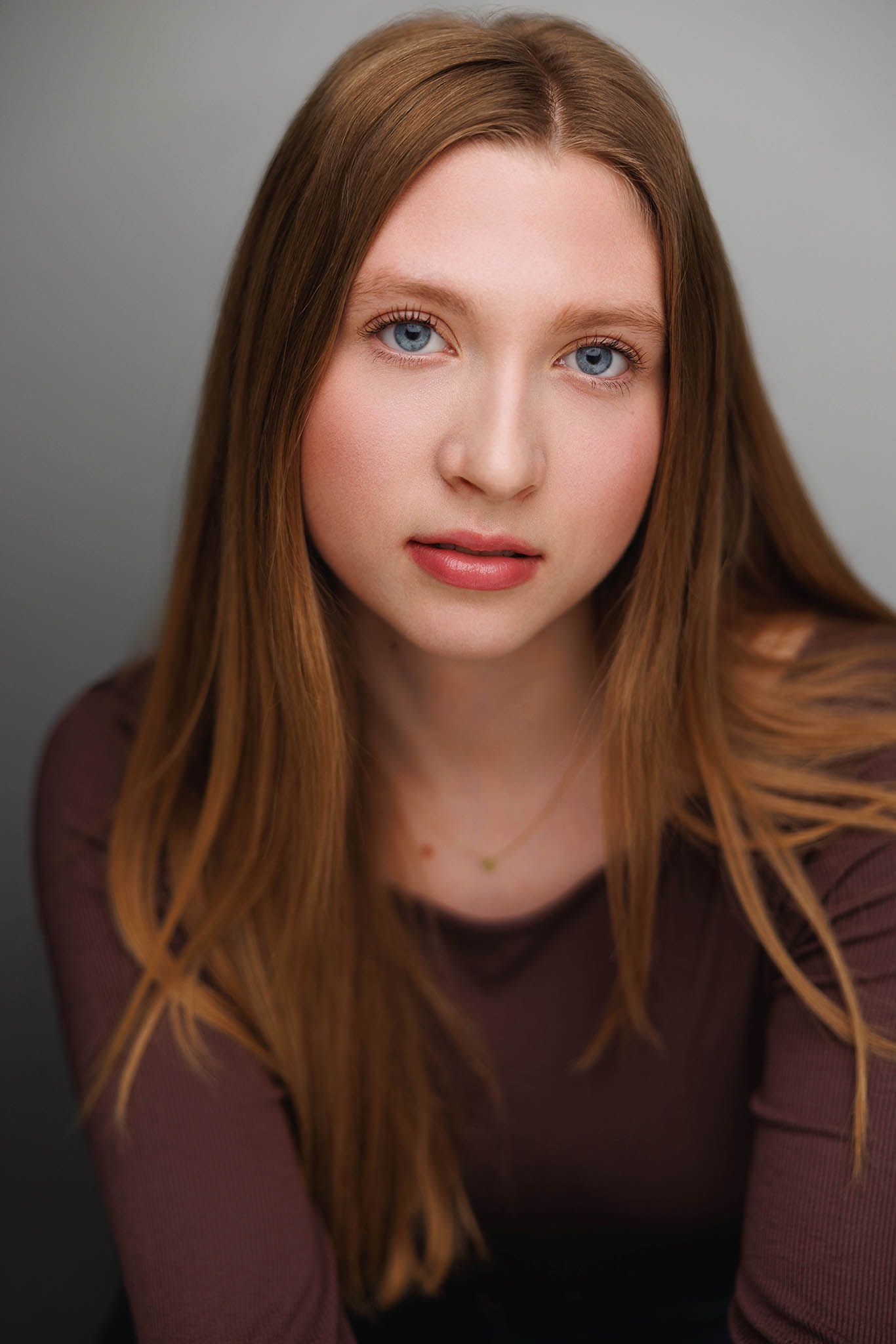 Portrait of a young woman with long auburn hair and striking blue eyes wearing a brown top against a gray background.