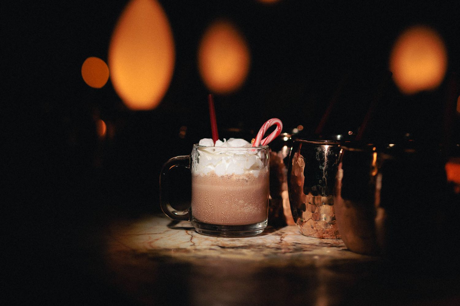 A festive holiday cocktail with whipped cream and candy cane garnish sits on a dark wooden table with bokeh lights in background.