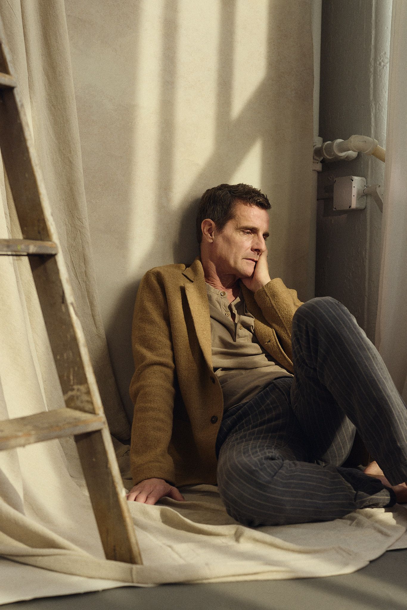 Man in tan blazer and striped trousers sits pensively on floor leaning against wall beside wooden ladder.