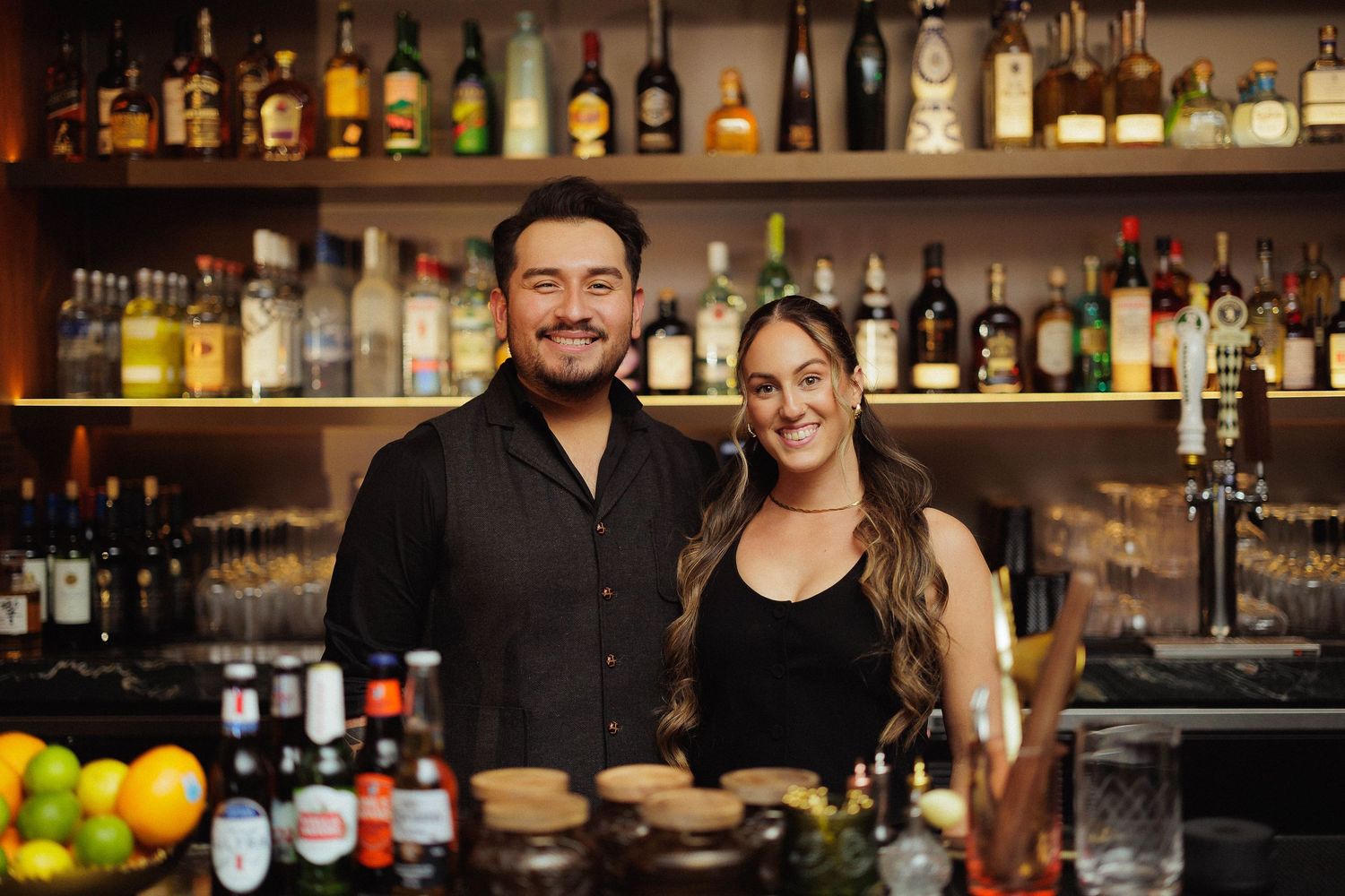 A well-stocked bar display with rows of liquor bottles illuminated on wooden shelves against a dark background.