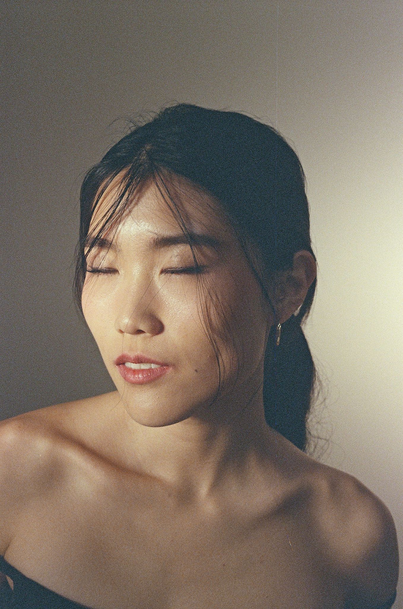 Young Asian woman with eyes closed, wearing gold earrings, bare shoulders, hair pulled back, soft studio lighting.