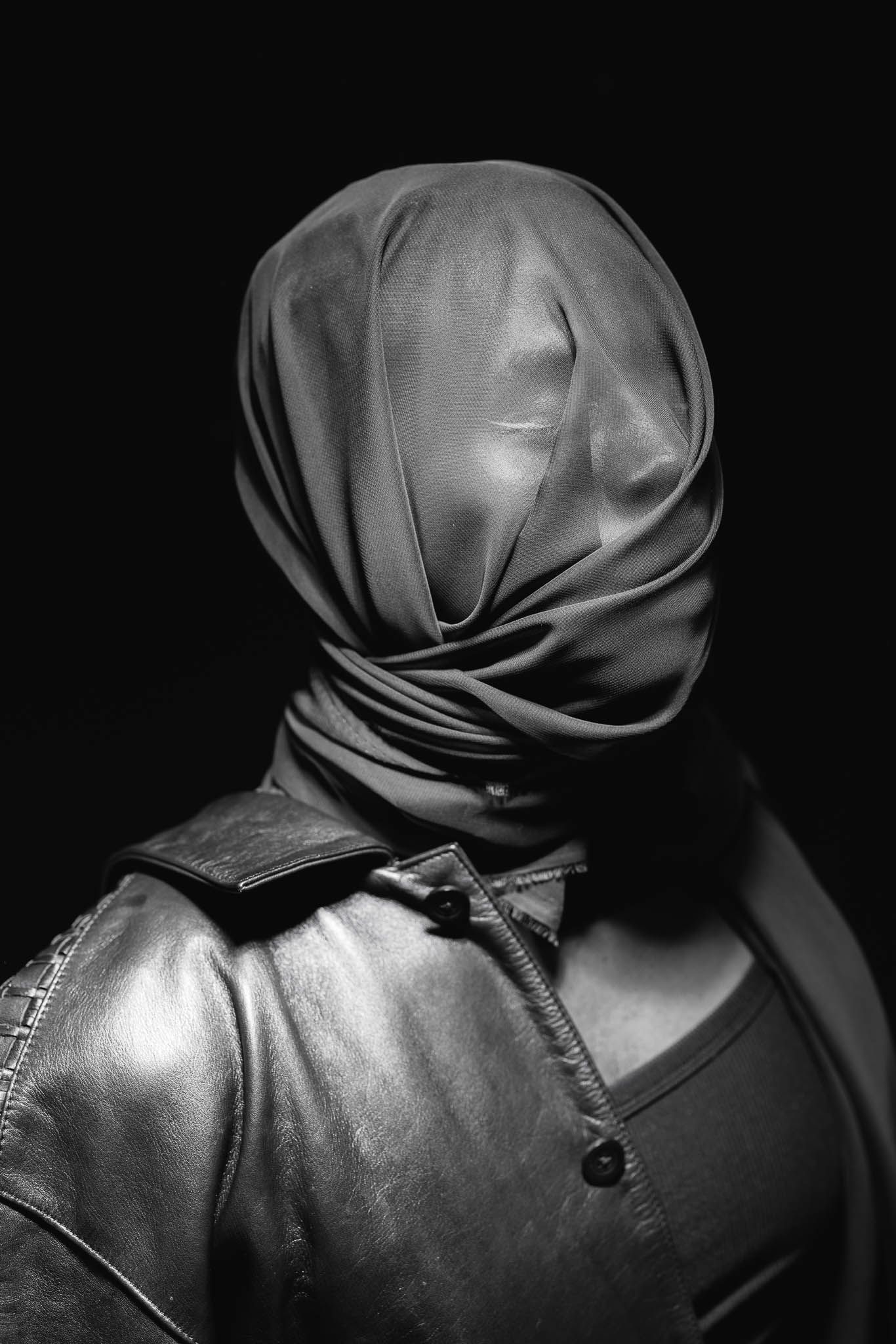 Dramatic black and white portrait of figure wearing wrapped fabric headpiece and leather jacket.