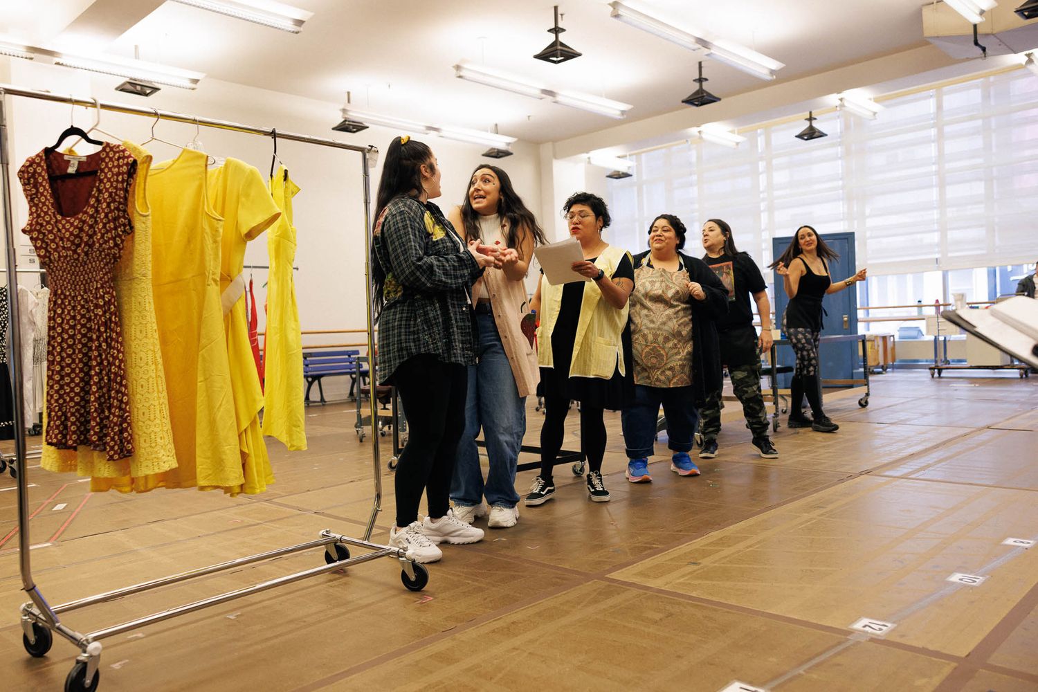 Theater group gathered around clothing rack during rehearsal in bright studio space.