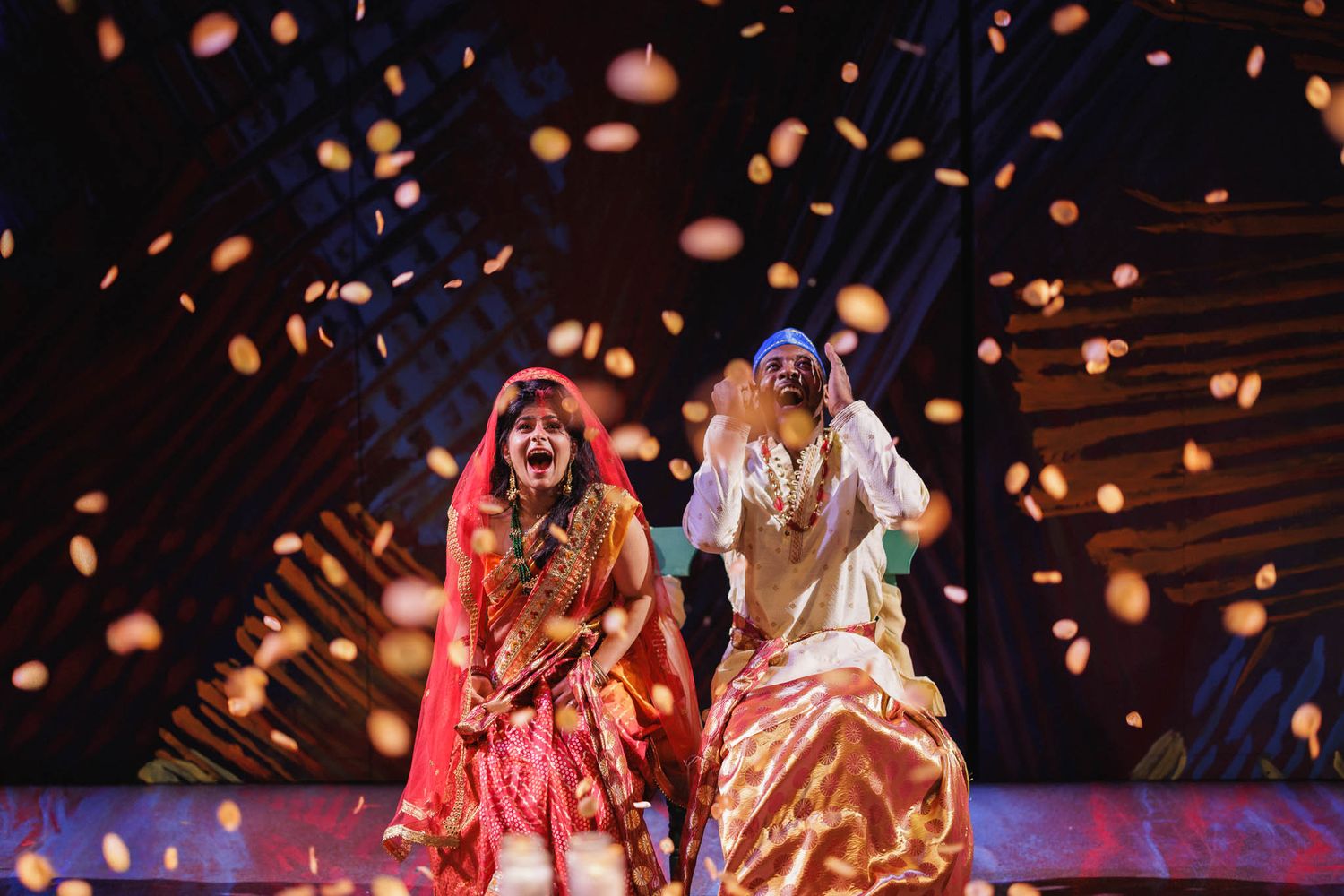 Performers in vibrant red and gold costumes dance on stage with glowing confetti falling around them in dramatic lighting.