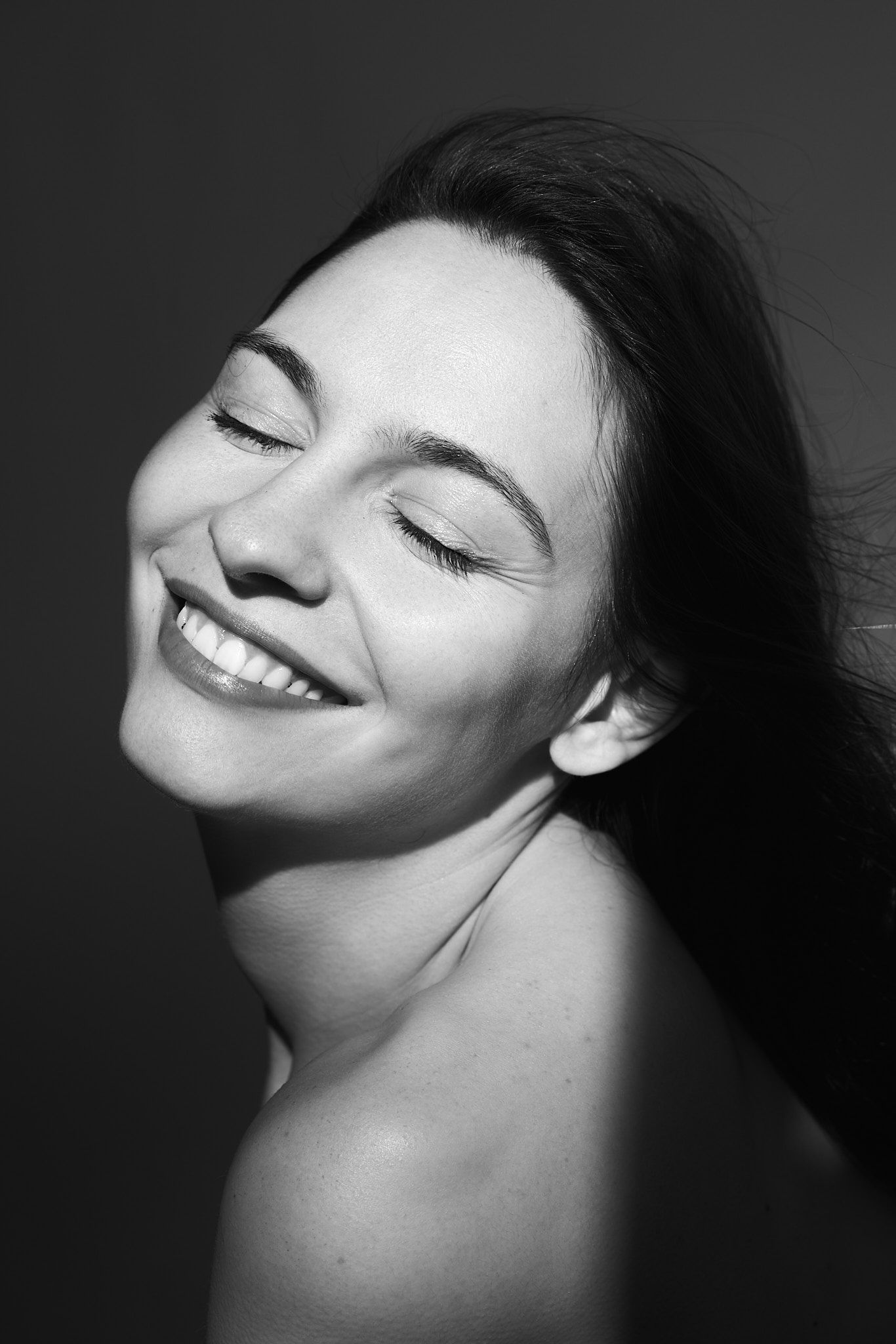 Black and white portrait of a smiling woman with closed eyes and bare shoulders against a dark background.