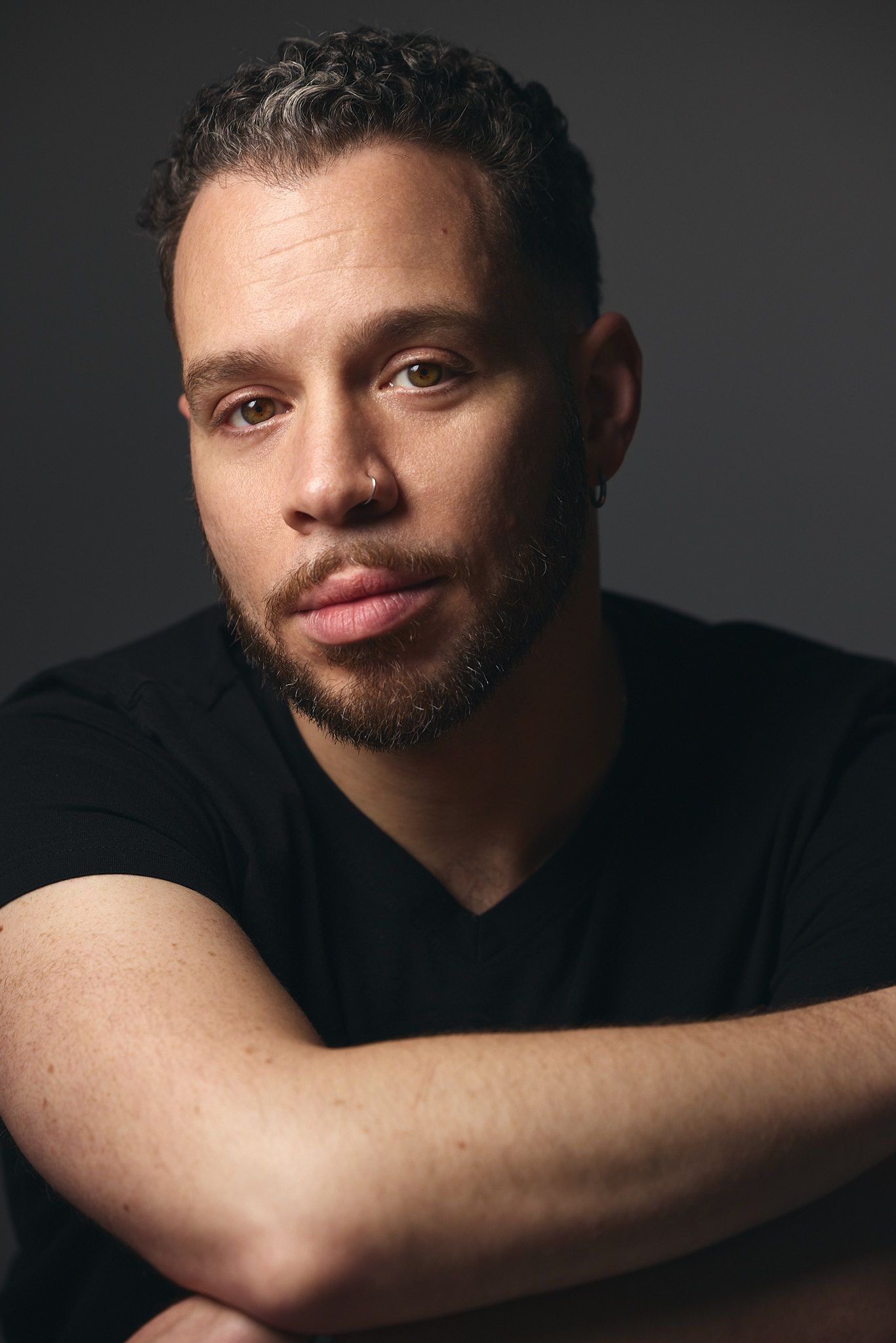 A bearded man in a black t-shirt poses with arms crossed against a dark gray studio background.