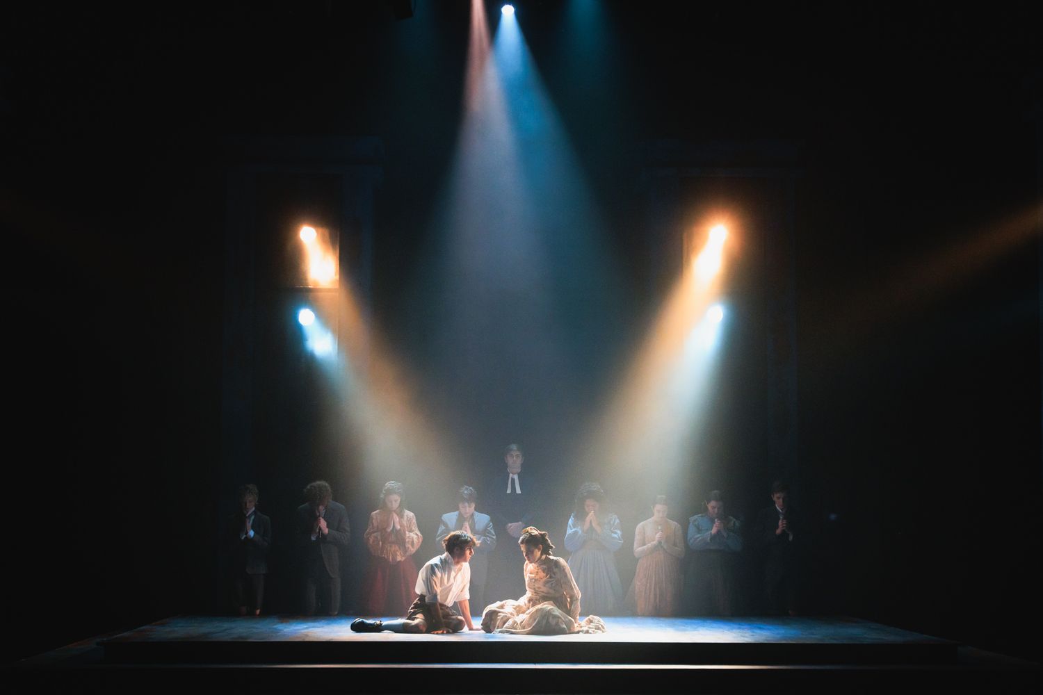 Two performers on stage under dramatic spotlights with ensemble cast in background.