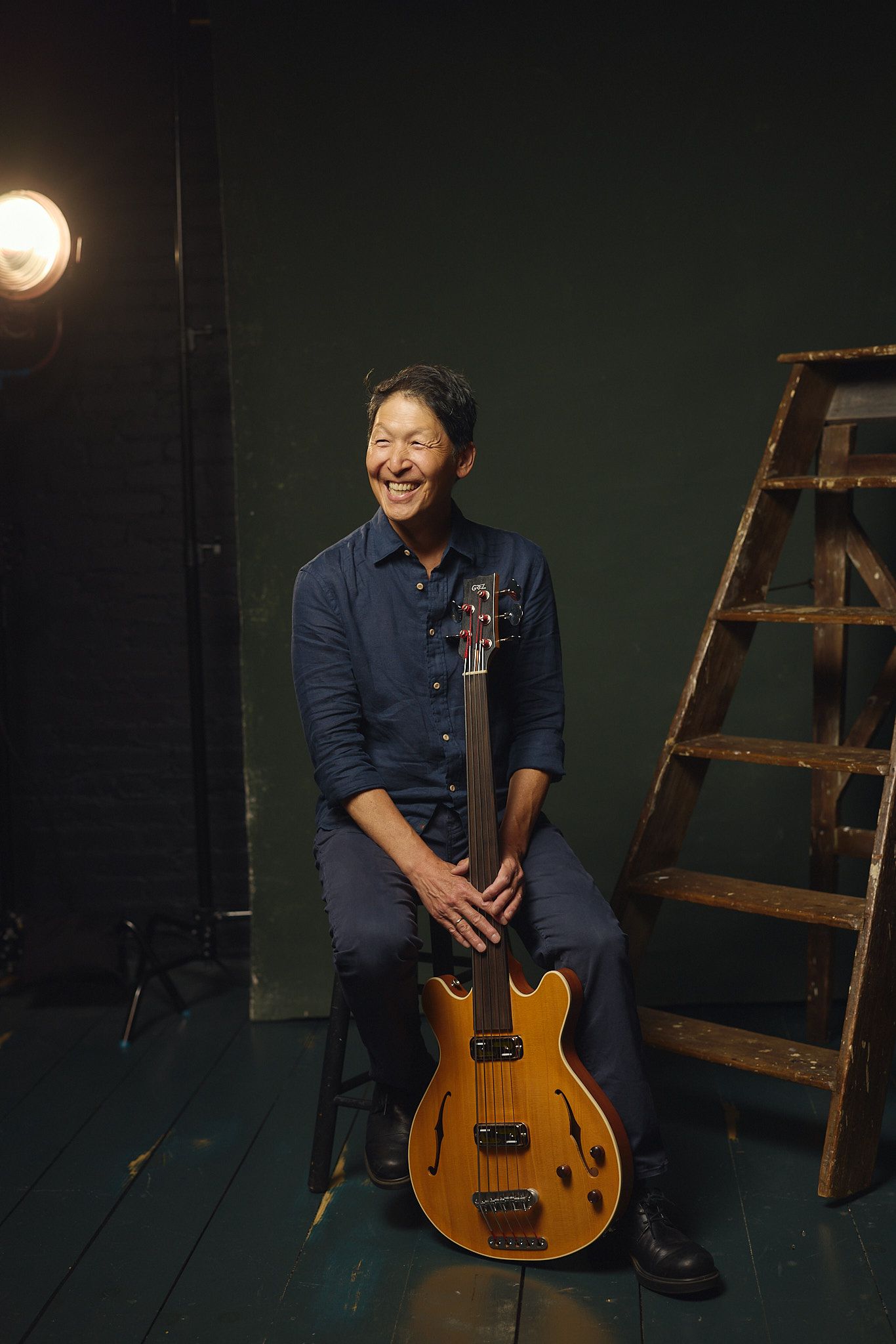 Smiling Asian man sitting with a vintage hollow-body electric guitar in a dark studio with a ladder and spotlight.