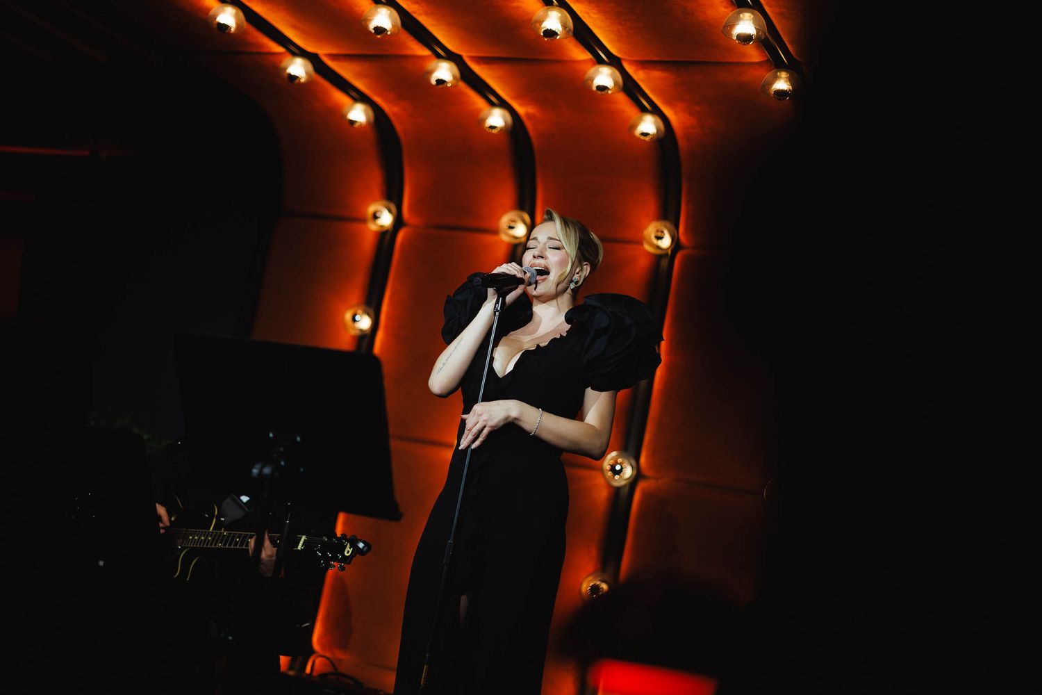 A performer in black clothing sings on stage under dramatic red lighting during a live performance.