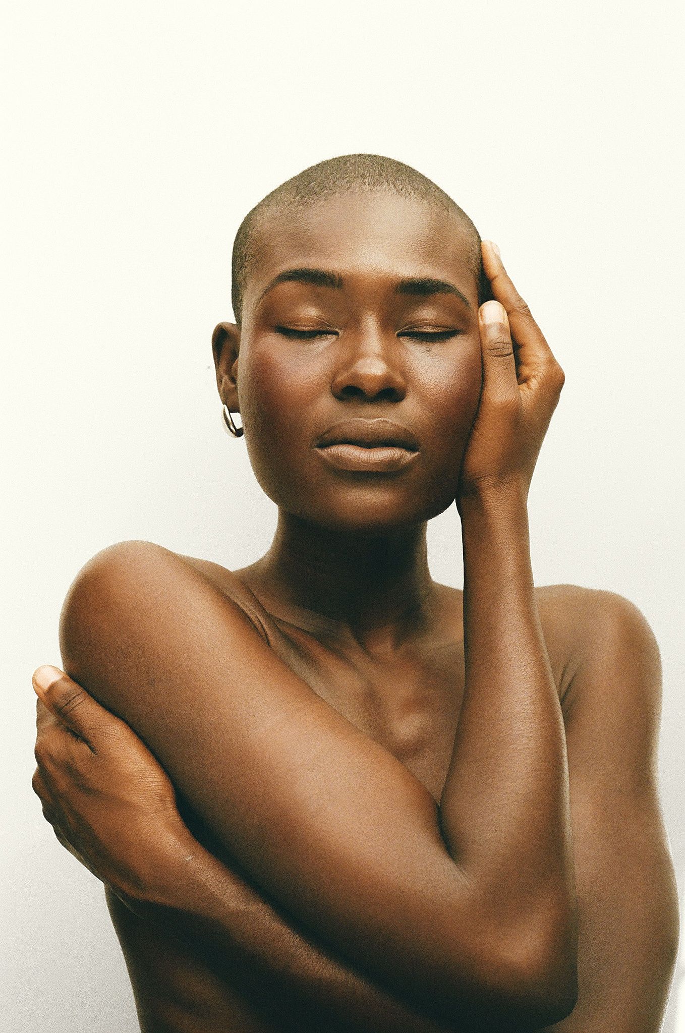 A young Black woman with a shaved head poses gracefully with closed eyes, touching her face against a white background.
