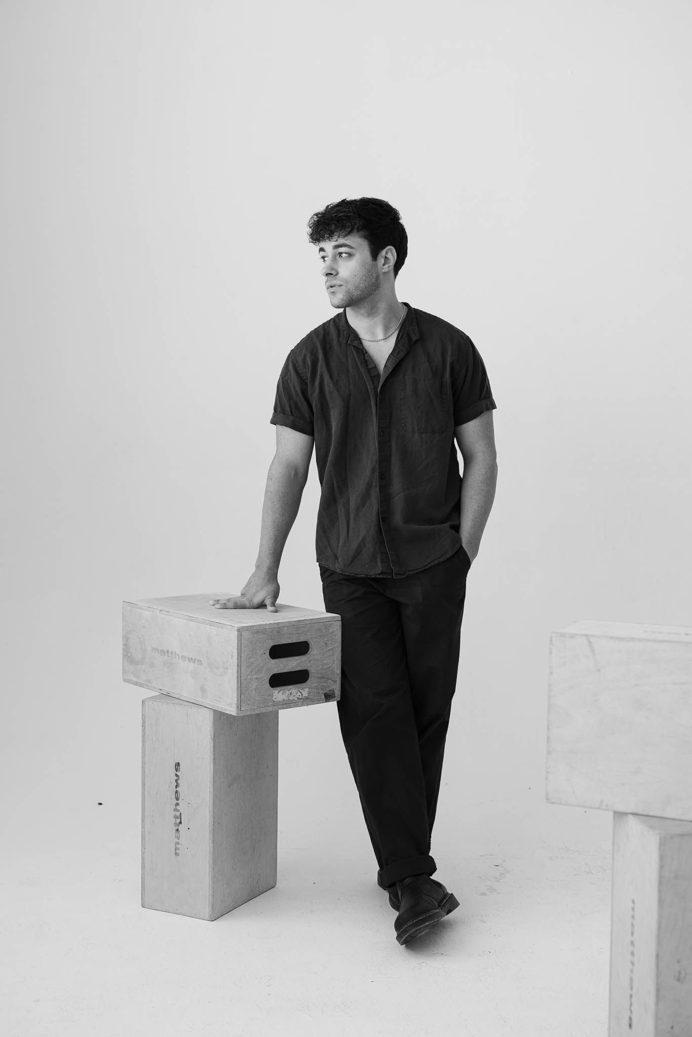 Black and white portrait of someone in casual attire leaning against white boxes in a minimalist studio setting.