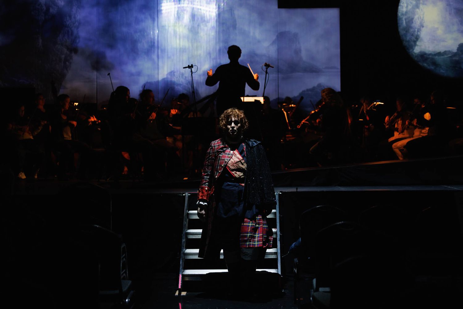 A conductor leads an orchestra on stage during a dramatic performance with moody blue lighting and fog effects.